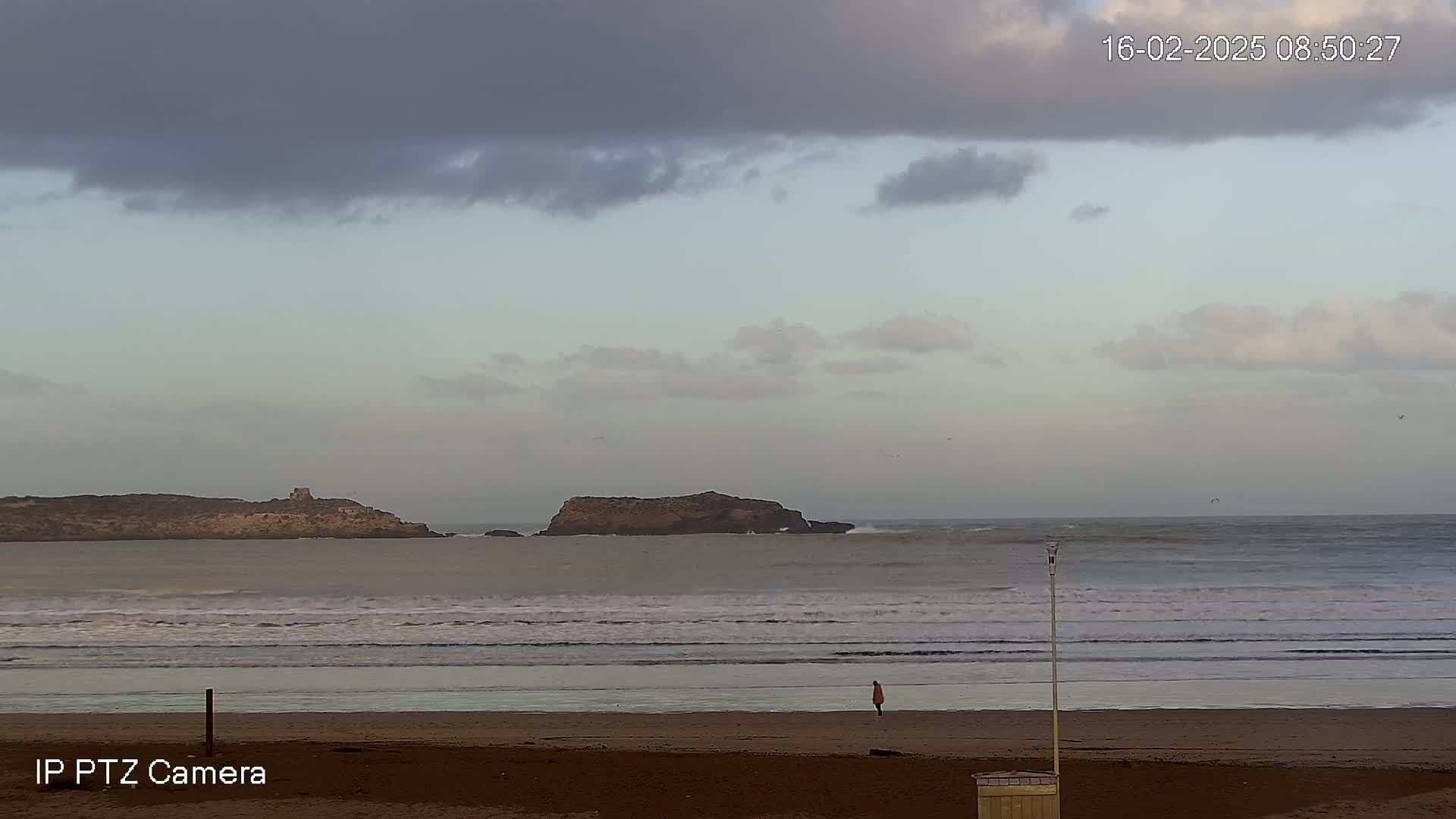 A person stands on a sandy beach overlooking a calm ocean with small waves, two rocky islands in the distance, and a cloudy sky.