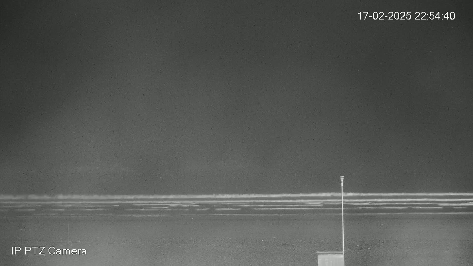 A nighttime, low-light view shows a dark beach with small waves lapping the shore under a mostly cloudy sky.
