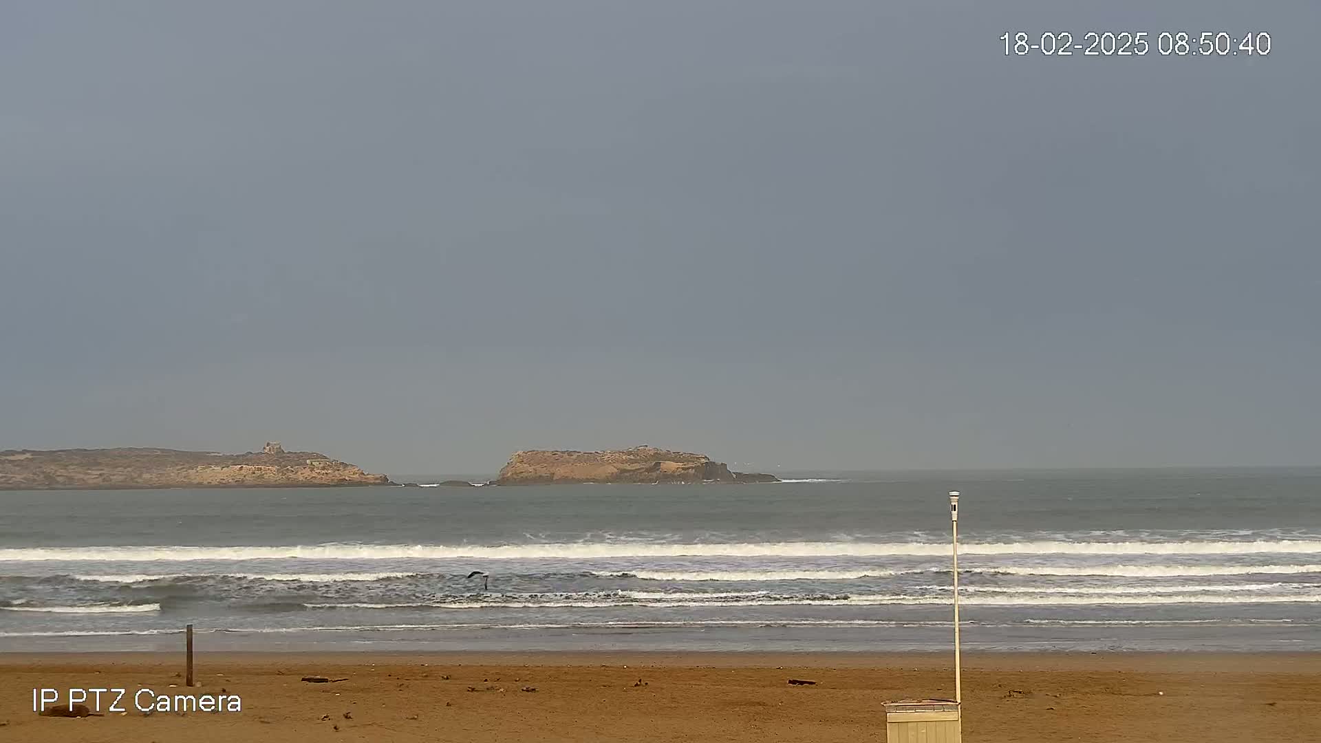 A sandy beach with gentle waves rolling in toward the shore under an overcast sky, with a small island visible in the distance.