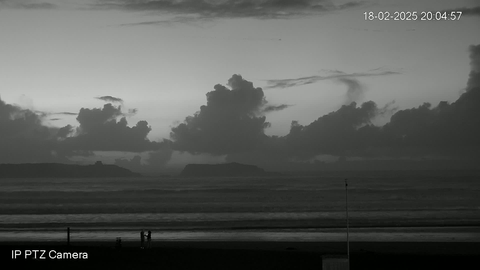 A grayscale image shows a dark, cloudy sky over a beach with a choppy ocean and a few people standing near the shoreline.
