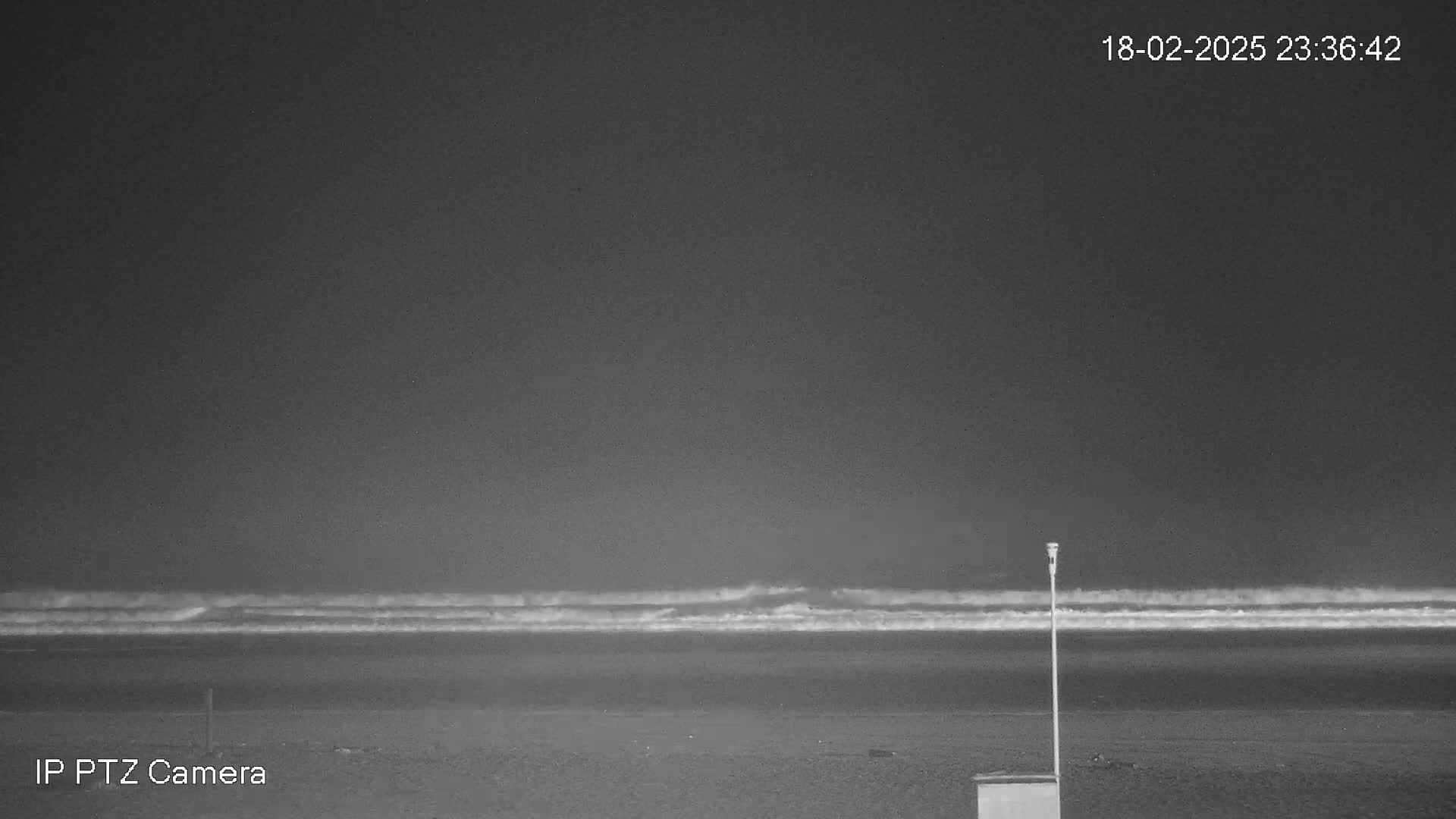 A dark, nighttime beach scene shows gently rolling waves under a mostly clear sky, with a single pole and a small box visible on the sand.