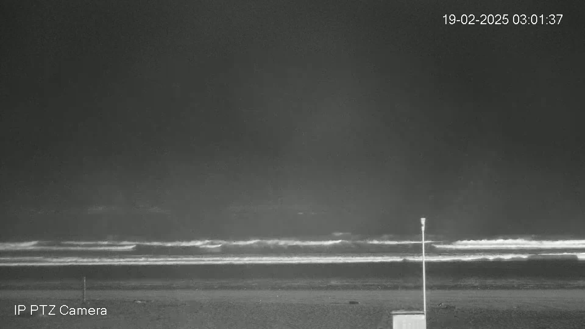 A dark, nighttime beach scene shows waves breaking on the shore under a cloudy sky.