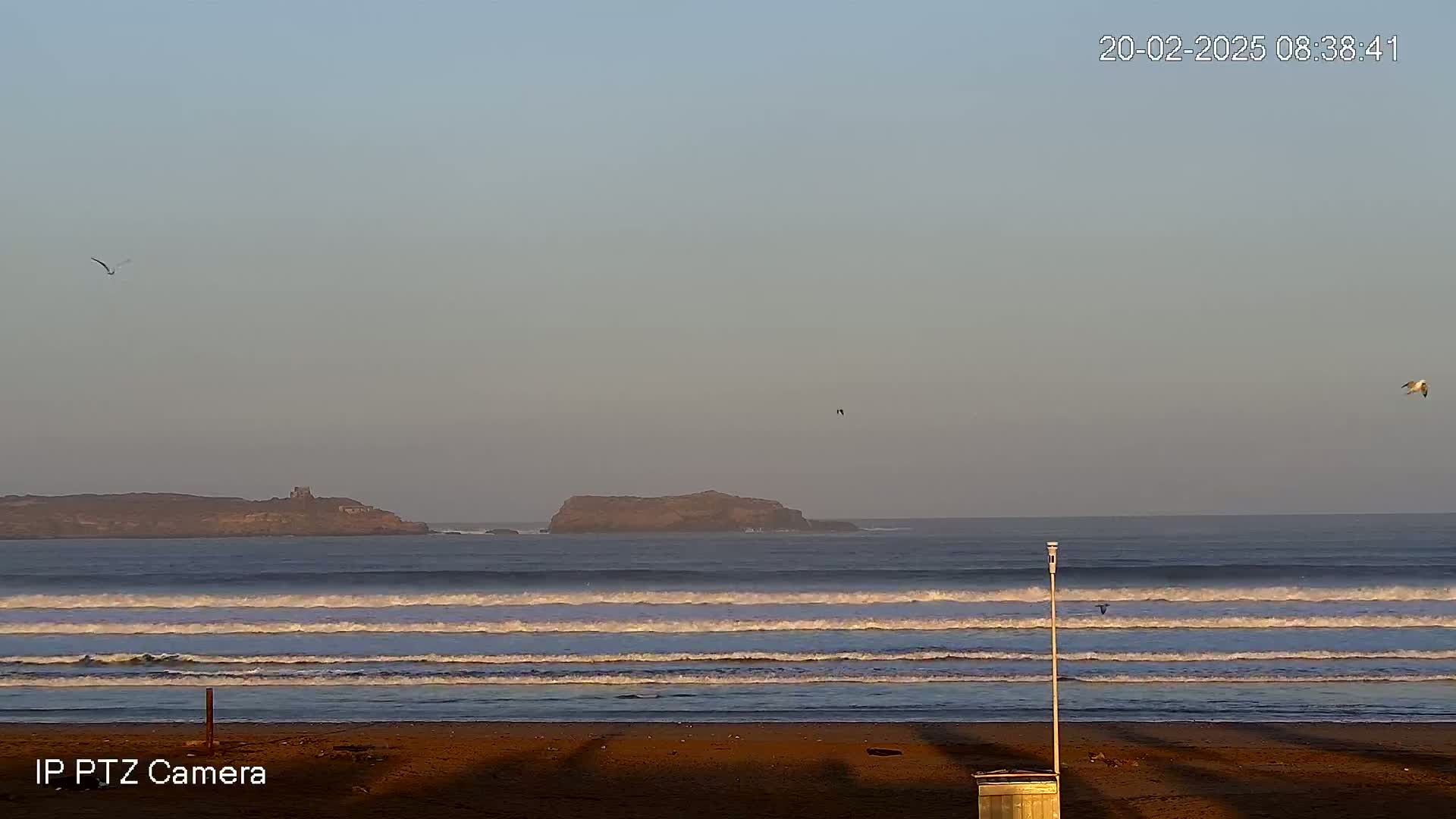 A sandy beach with rolling waves and two small islands in the distance under a clear, pale sky.