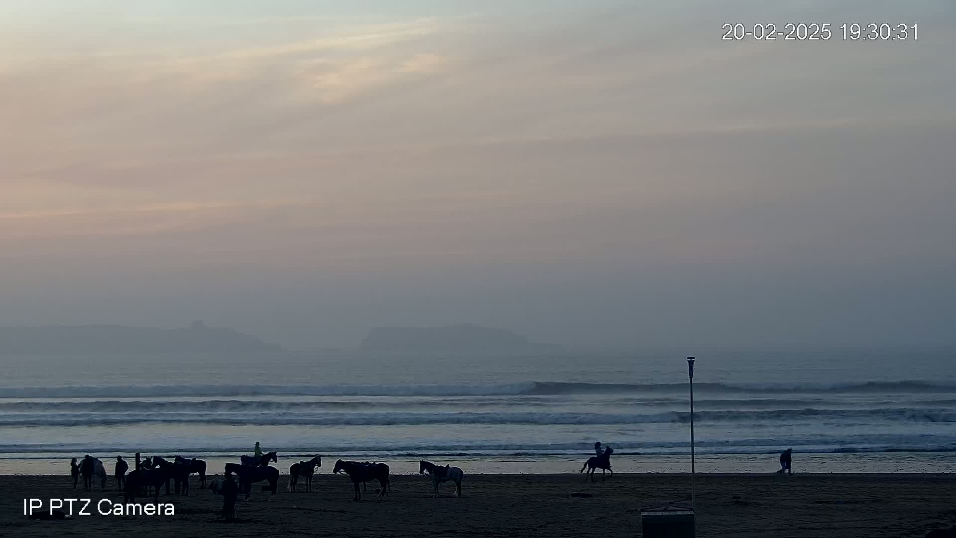 Several people ride horses on a hazy beach at sunset, with a distant island visible through the mist.