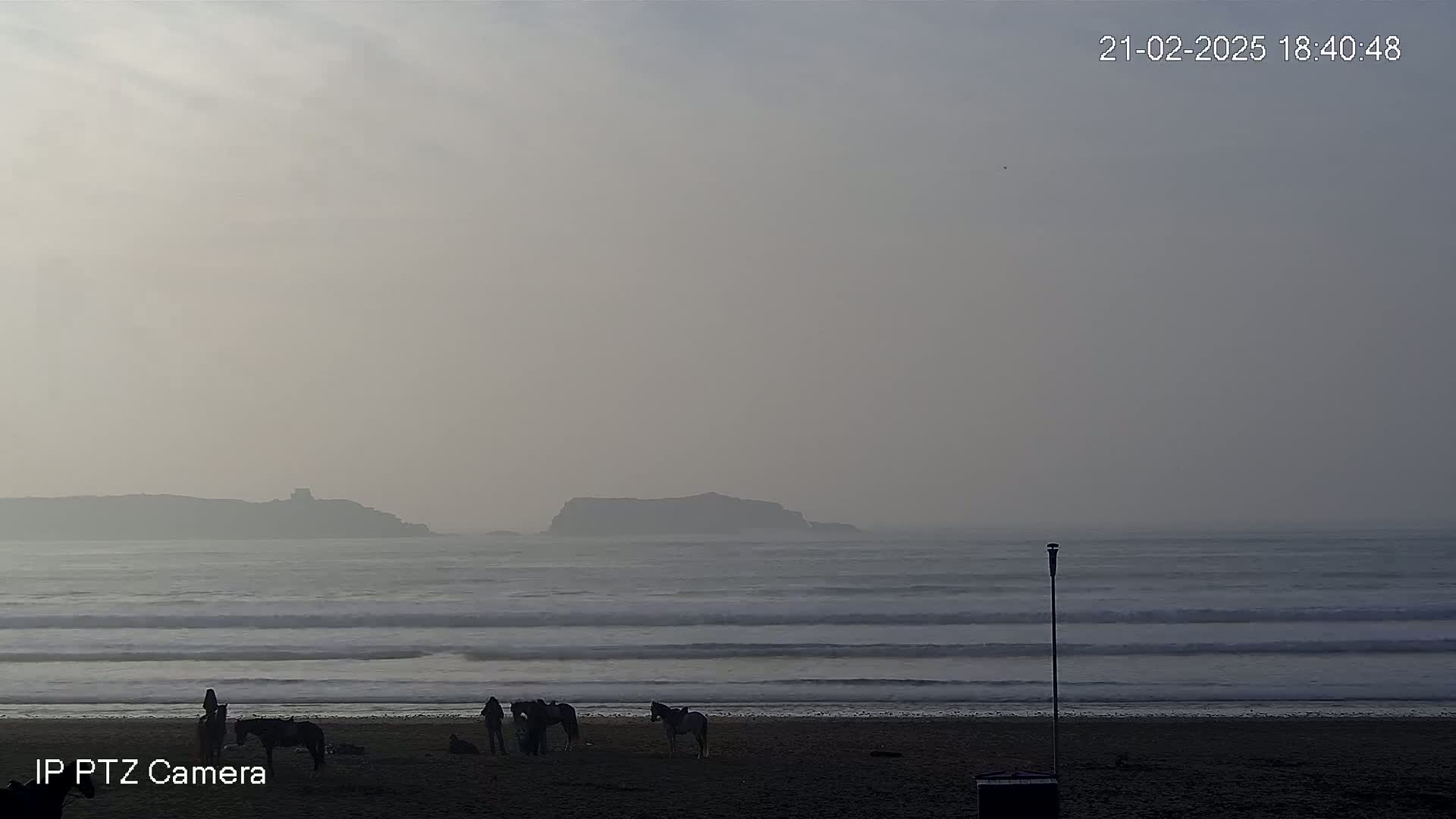 A hazy beach scene shows several people with horses on the sand, with waves rolling in toward a distant, low-lying island.