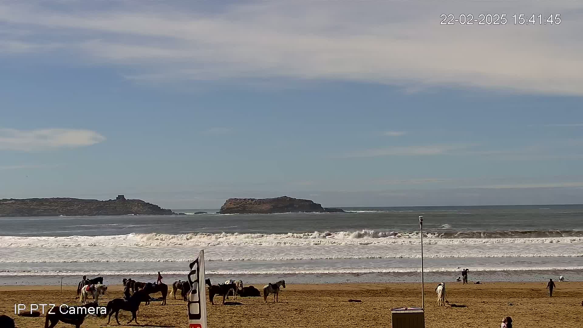A sandy beach with several horses and riders, under a partly cloudy sky, with moderate waves breaking in the ocean beyond.
