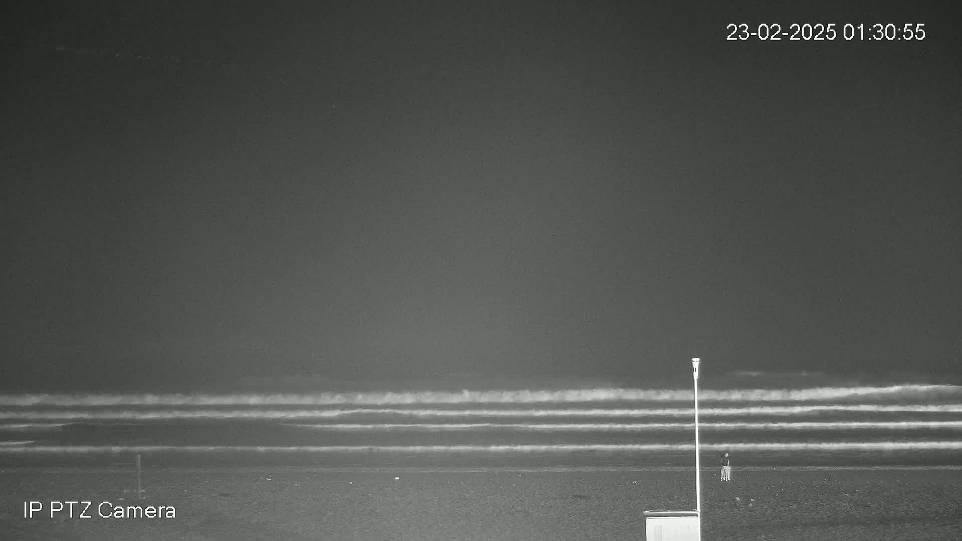 A dark, nighttime beach scene shows a person standing near a pole and a small structure, with gently rolling waves visible in the distance under a cloudy sky.