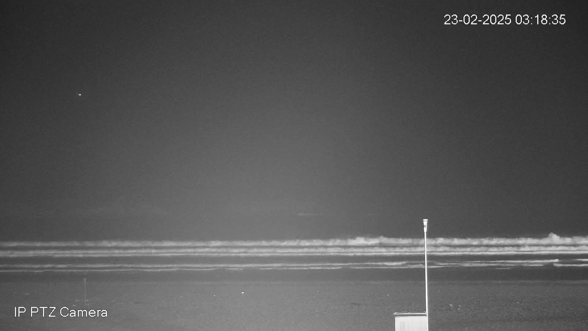 A dark, nighttime beach scene shows gently rolling waves under a clear sky with a single bright light visible in the distance.