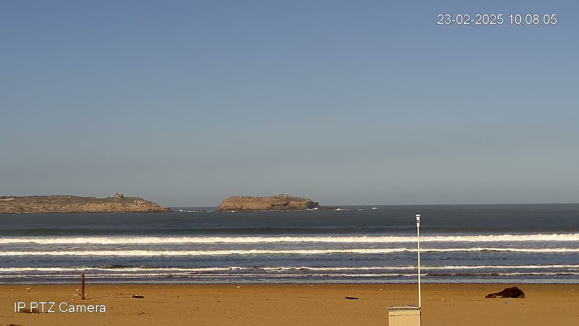 Under a clear blue sky, a sandy beach with gentle waves rolls towards two small islands in the distance.