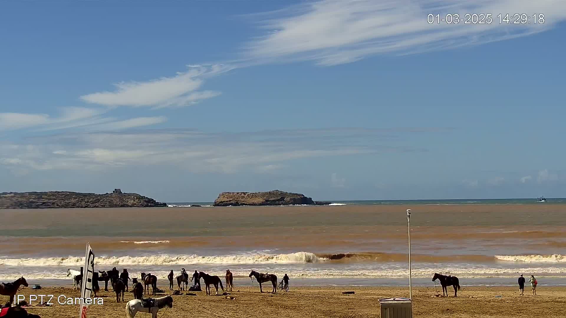 A sandy beach with several horses and riders, under a mostly clear blue sky with thin clouds, features slightly choppy brown water with small waves breaking on the shore.