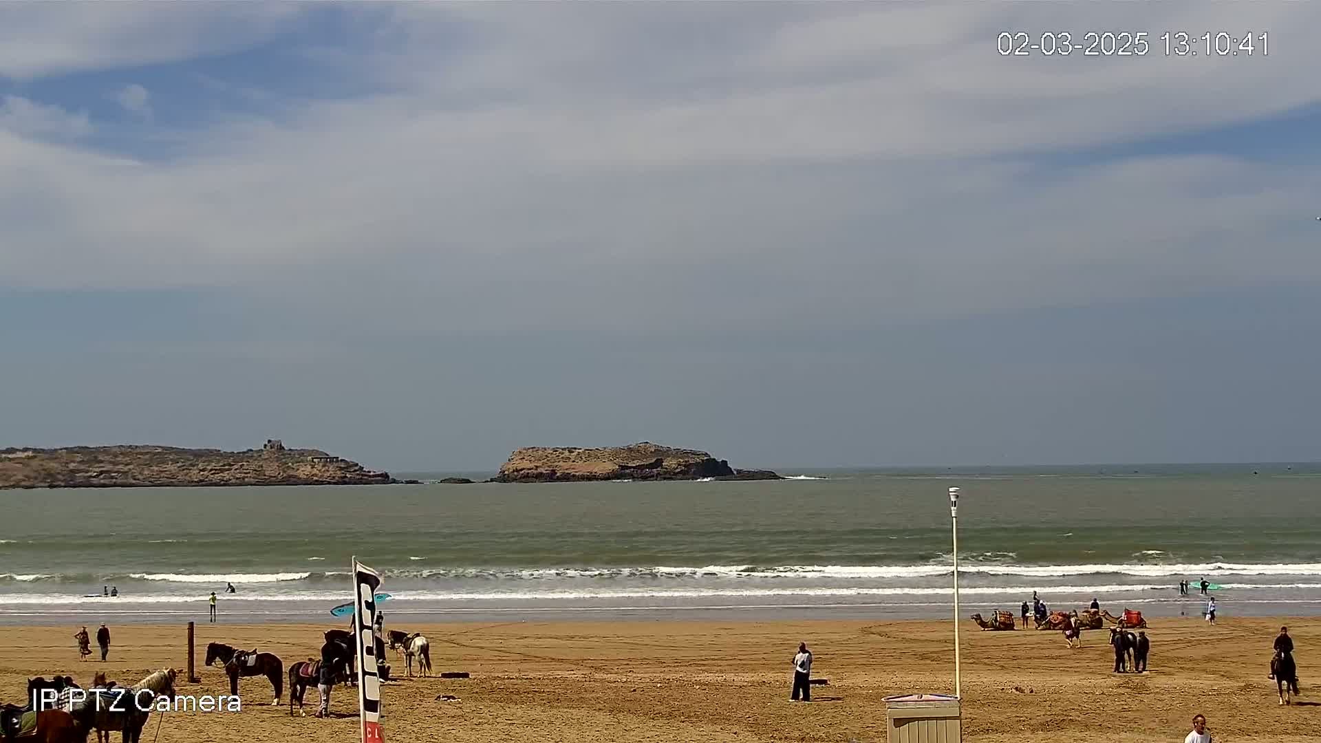 A sandy beach with several people, horses, and camels under a partly cloudy sky, features gentle waves and a small island in the distance.