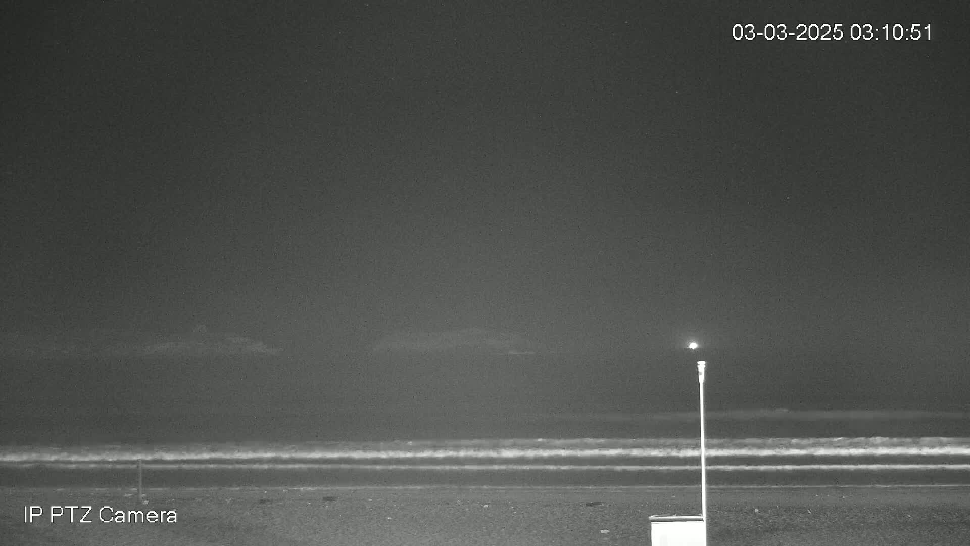 A dark, nighttime beach scene shows gently lapping waves under a clear sky, with a single light post standing near a small, square object on the sand.