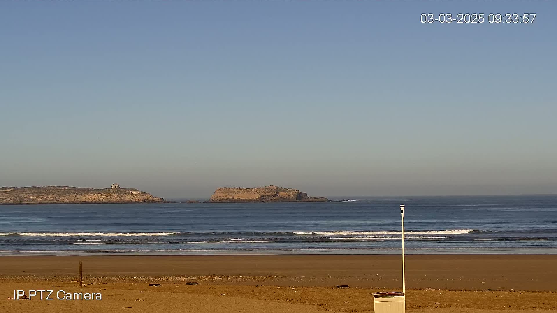 A sandy beach with gentle waves rolls onto the shore under a clear, bright sky, with two small islands visible in the distance.