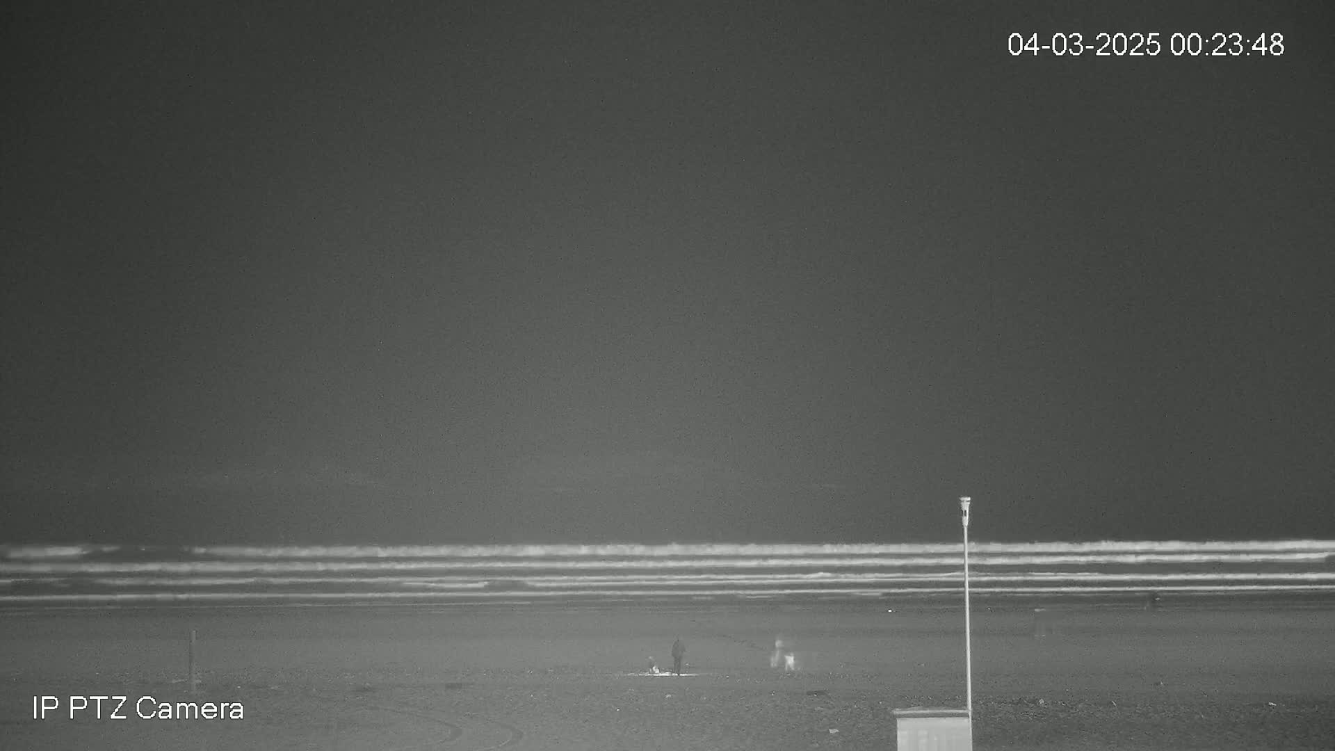 A nighttime beach scene shows a dark sky, gently rolling waves, and a few figures on the sand.