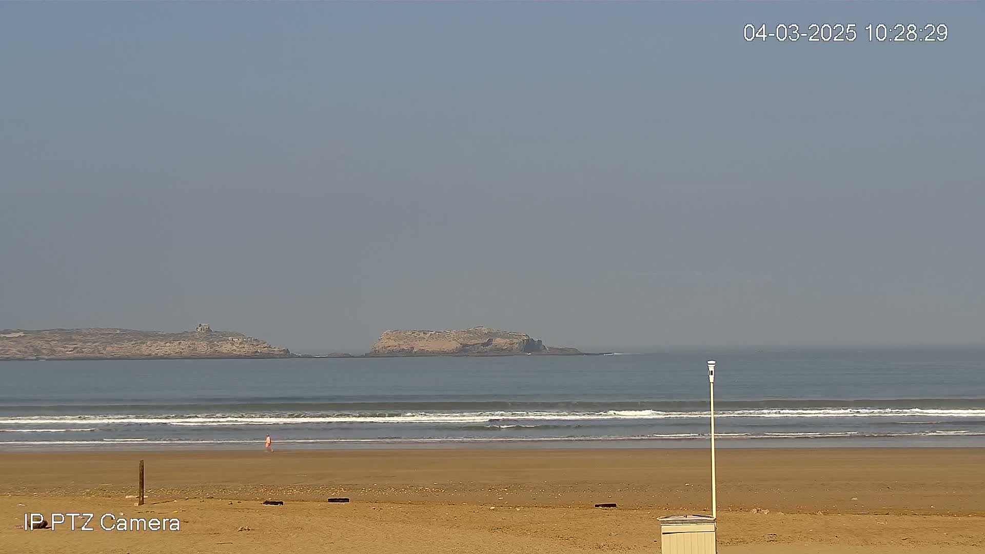 A sandy beach with gentle waves laps the shore, under a hazy sky, with two distant islands visible on the horizon.