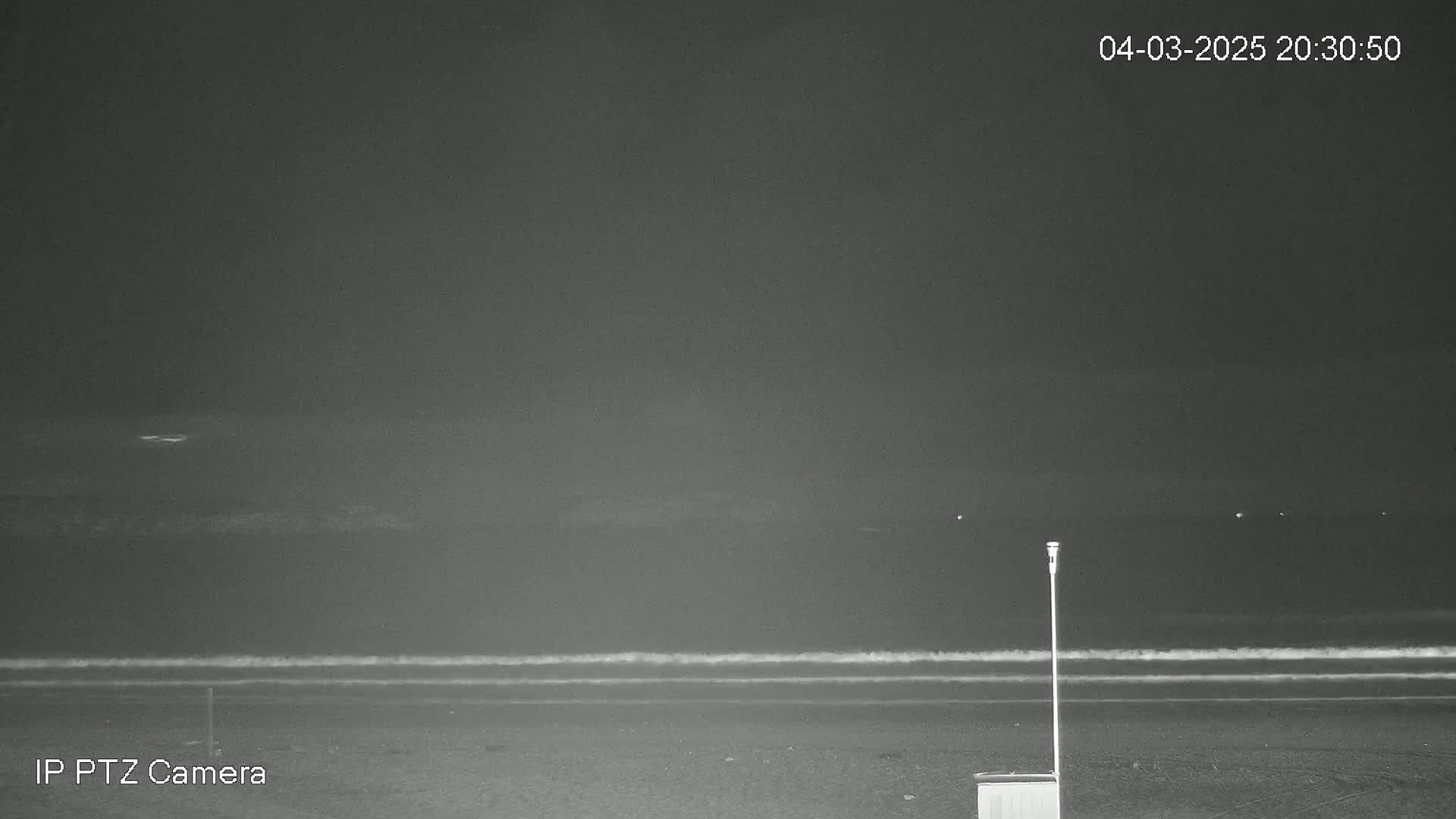 A dark nighttime beach scene shows a calm ocean with small waves under a mostly clear sky, and a single tall pole stands on the sand near a small box-like structure.