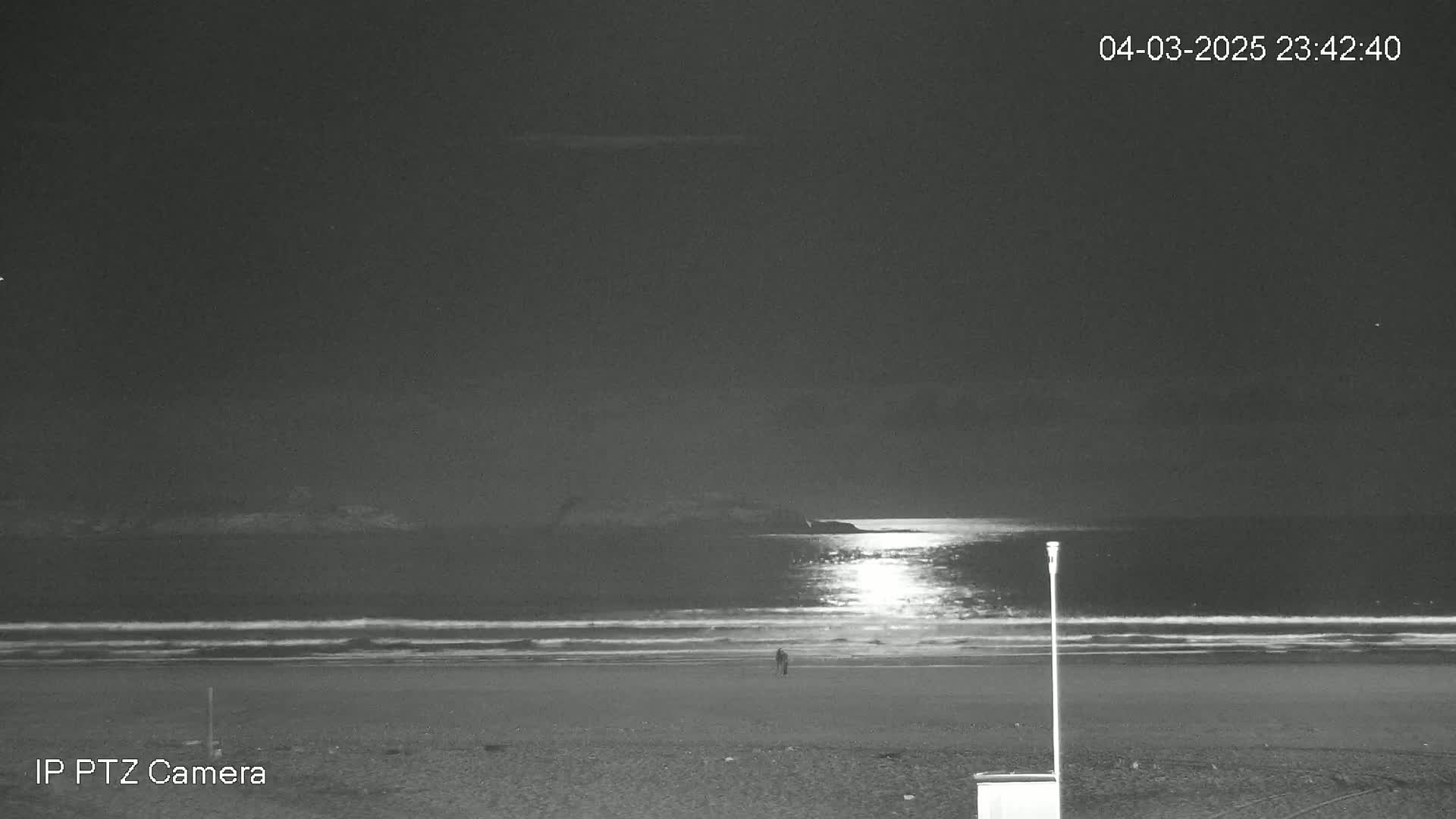 A dark, moonlit beach at night shows a single person walking near the water's edge.