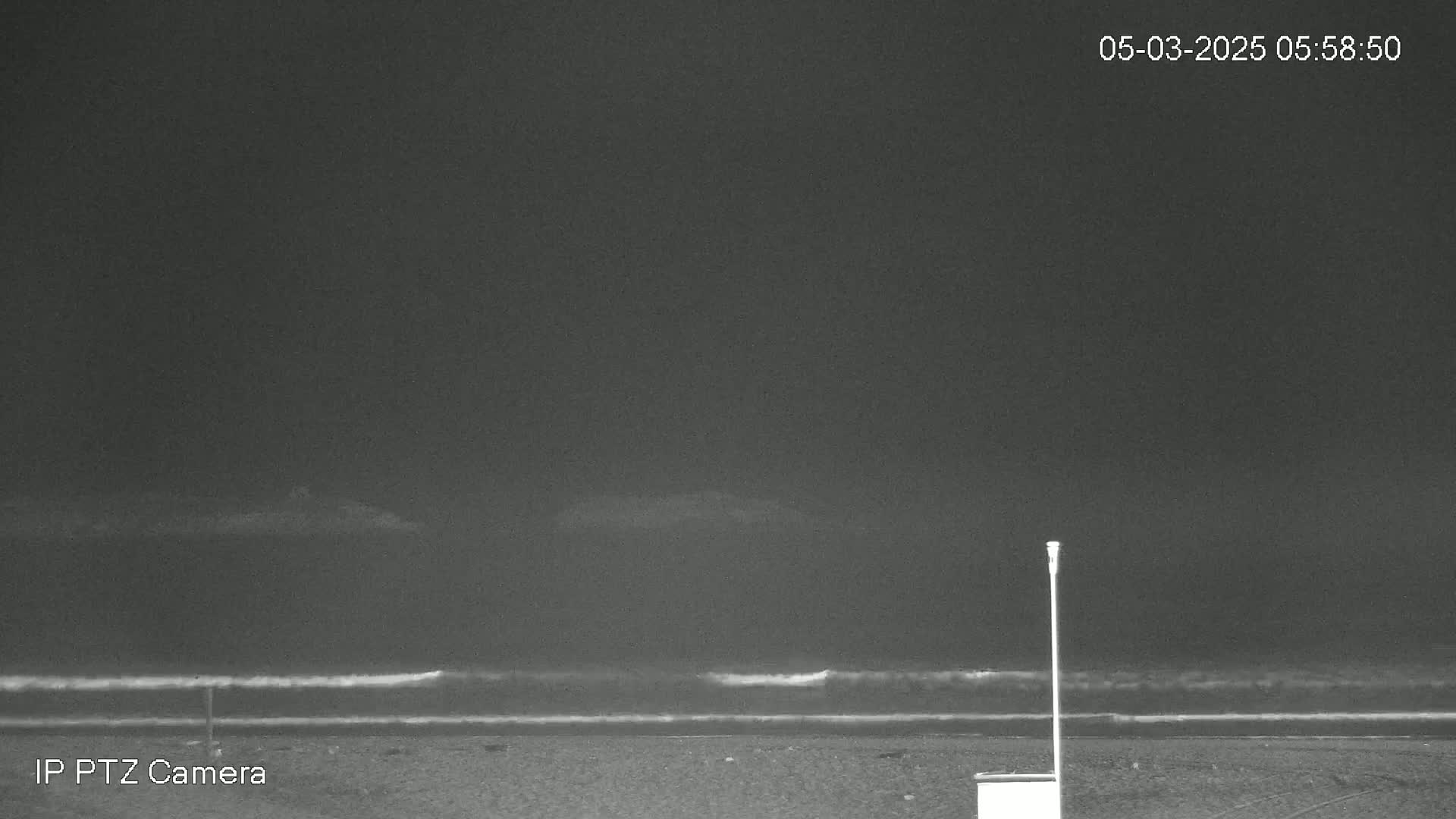 A dark, nighttime beach scene shows gentle waves lapping at the shore under a mostly clear, dark sky.