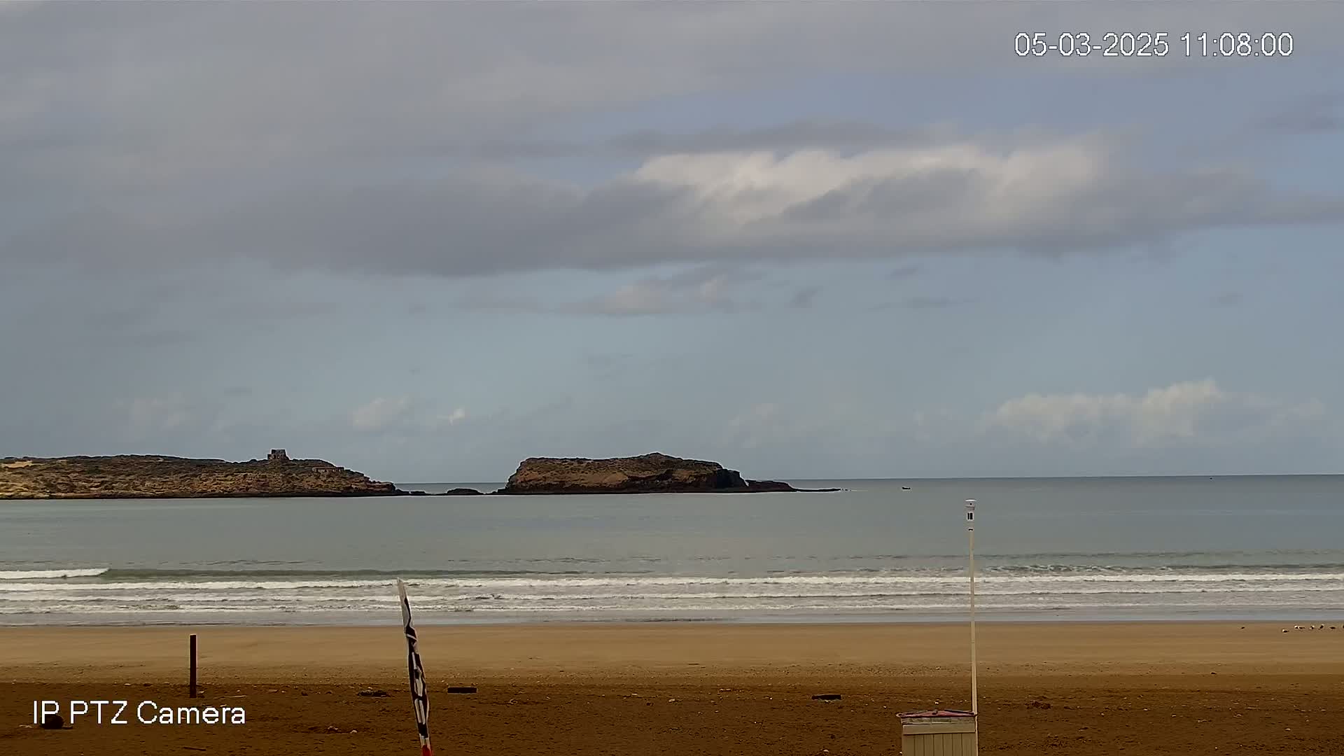 A sandy beach with small waves under a mostly cloudy sky shows two small islands in the distance.