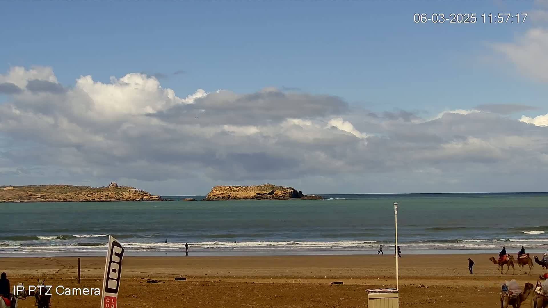 A sandy beach with people and camels is visible, with a calm ocean featuring small waves and two small islands in the distance under a partly cloudy sky.