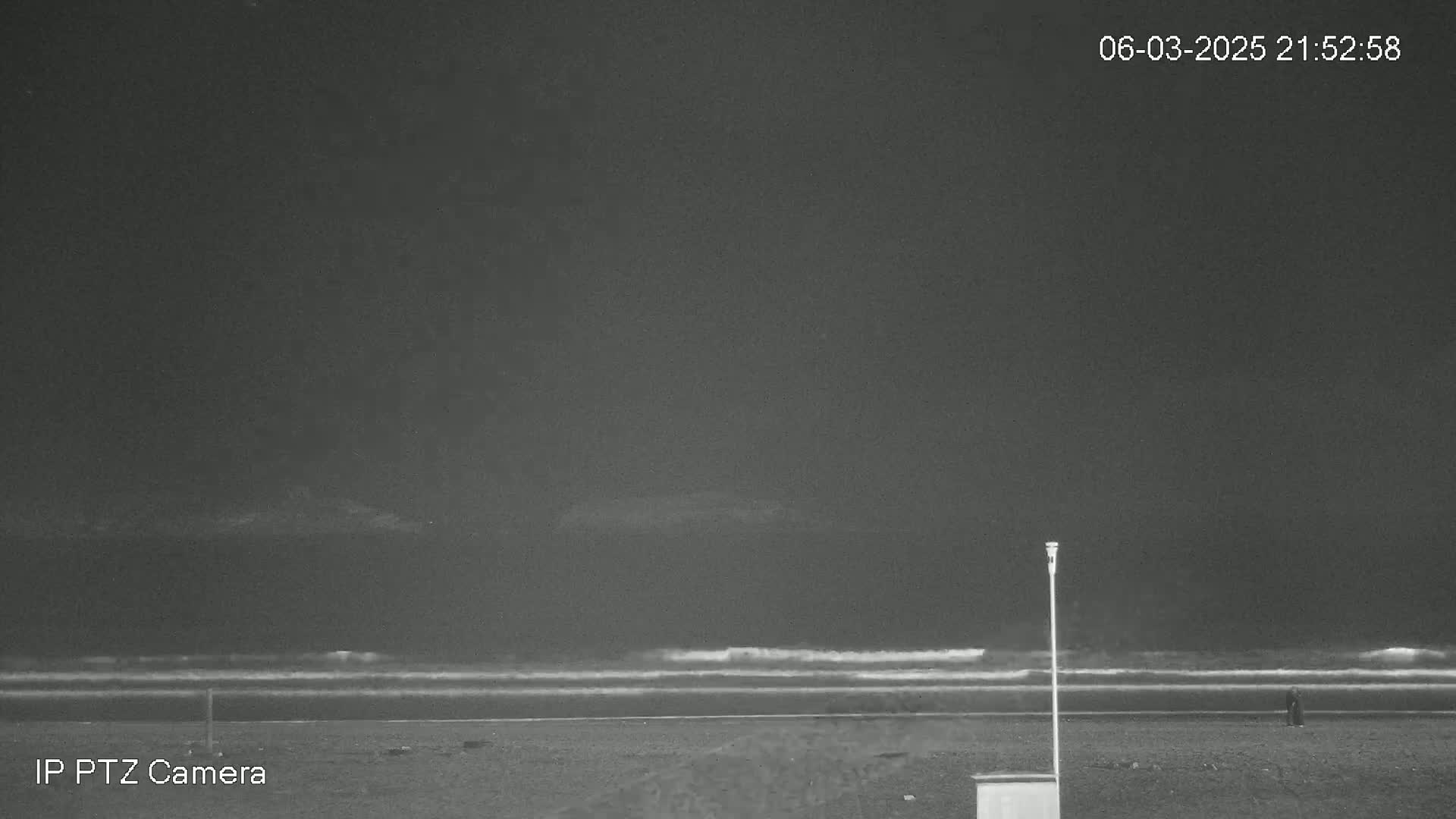 A nighttime beach scene shows a dark ocean with small waves, a dark sandy shore, and a single person in the distance under a cloudy sky.