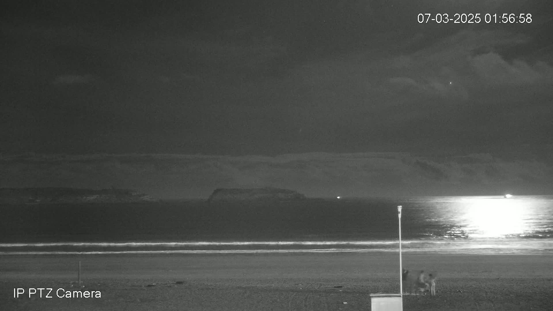 A dark, nighttime beach scene shows a calm ocean reflecting light, with a few people standing near the water's edge under a cloudy sky.