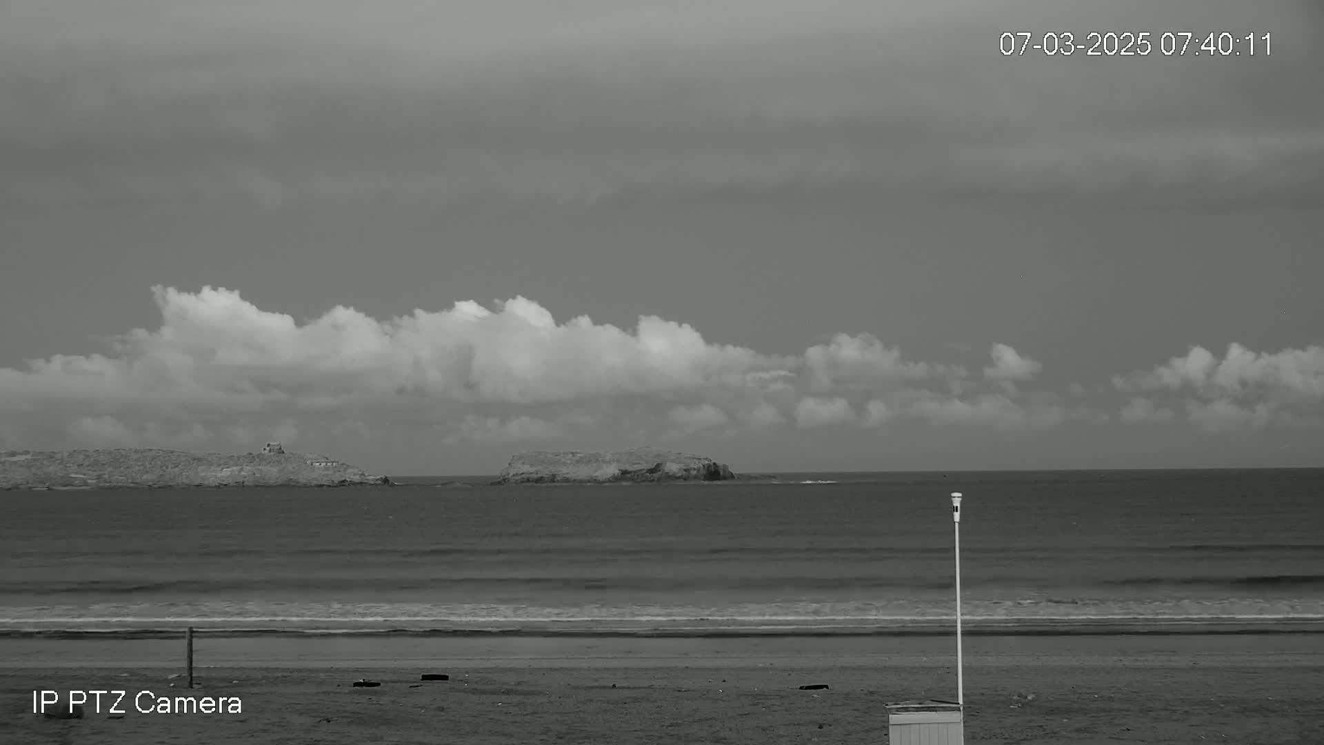 A grayscale image shows a beach with calm waves under a mostly cloudy sky, and two small islands visible in the distance.
