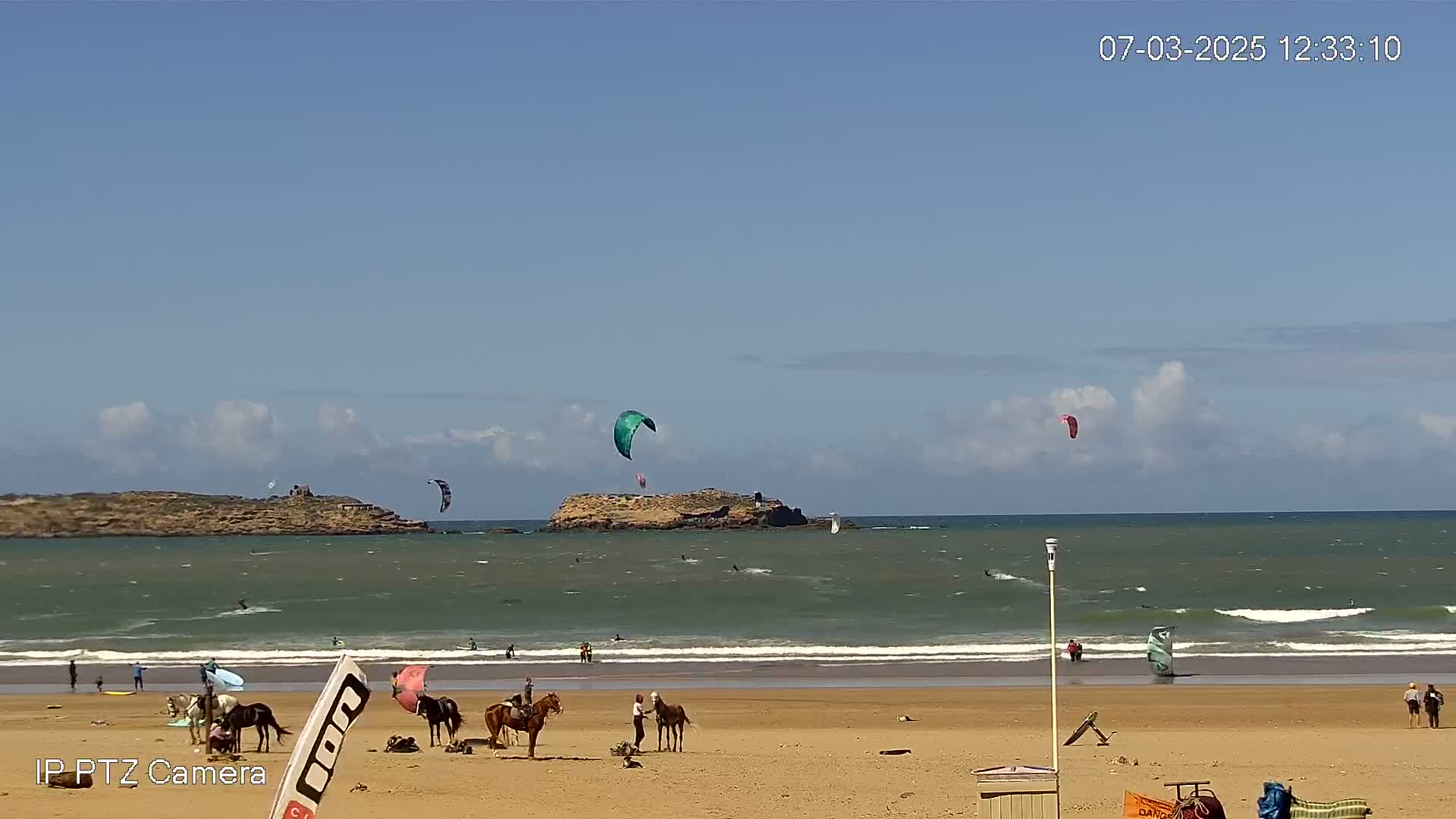 A sandy beach with several horses and riders, kite surfers in the ocean, and a small island in the distance under a mostly sunny sky.