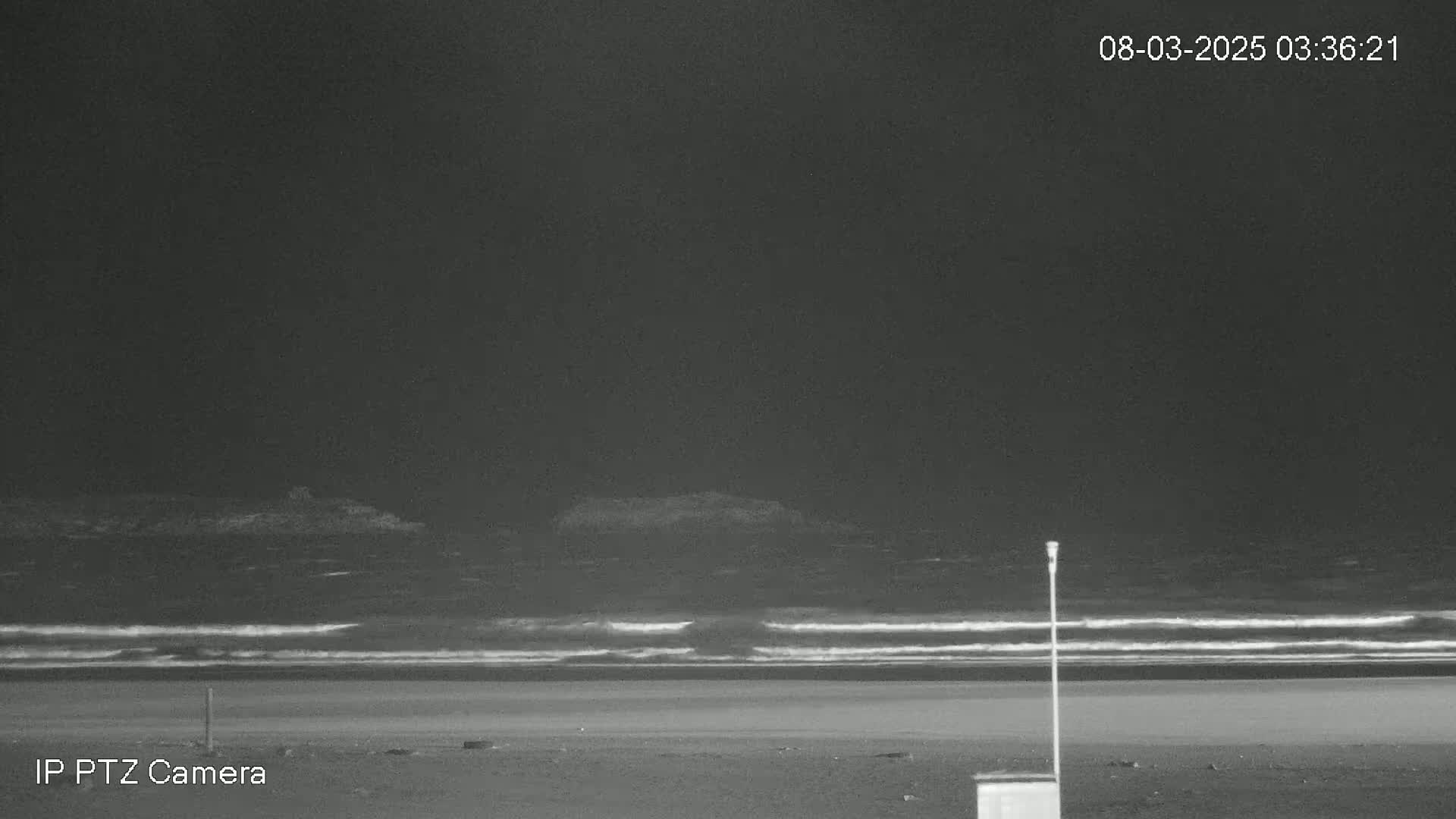 A dark, nighttime beach scene shows a calm ocean with small waves lapping the shore.