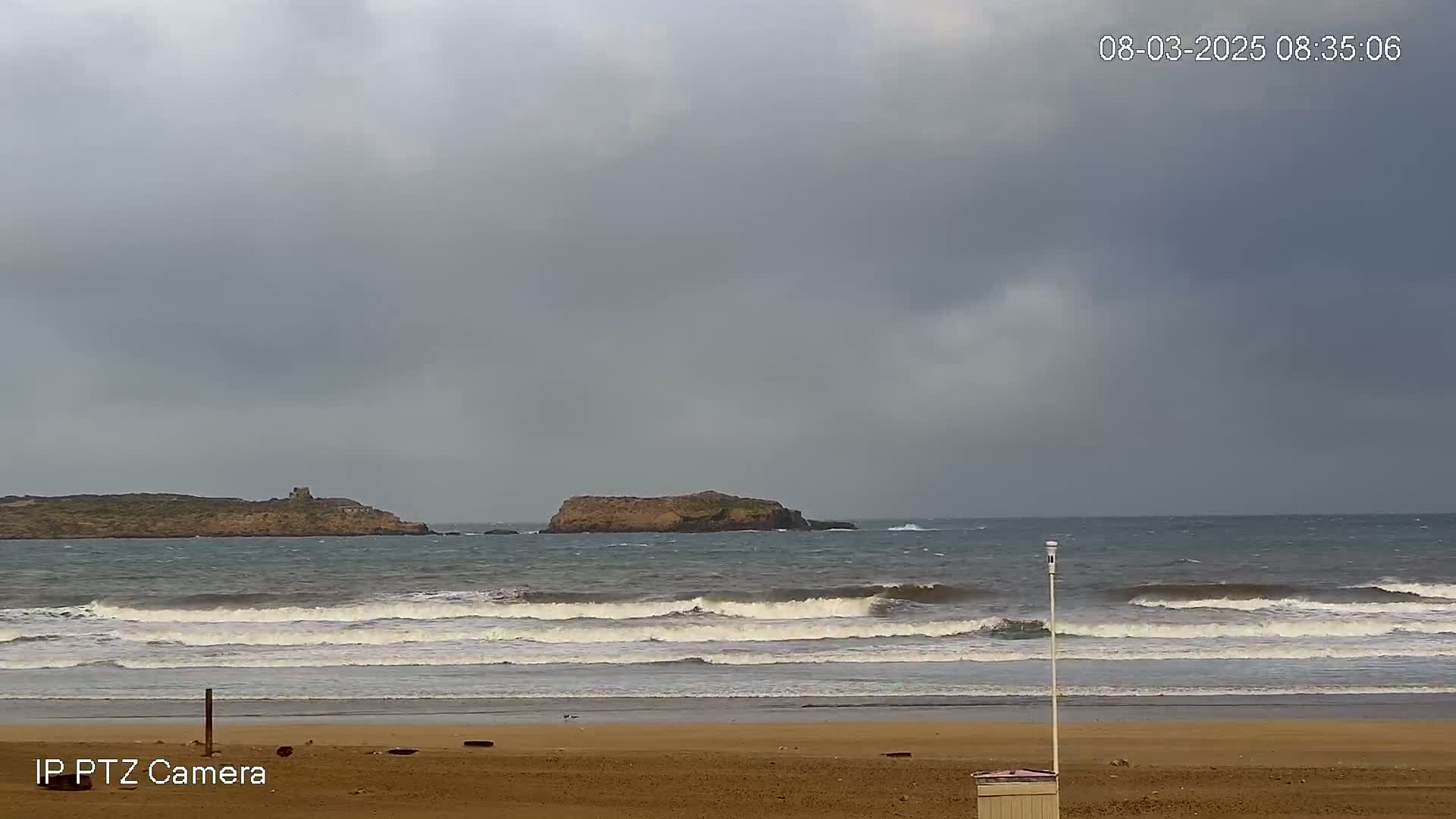 A sandy beach with moderate waves under a cloudy sky shows small islands in the distance.