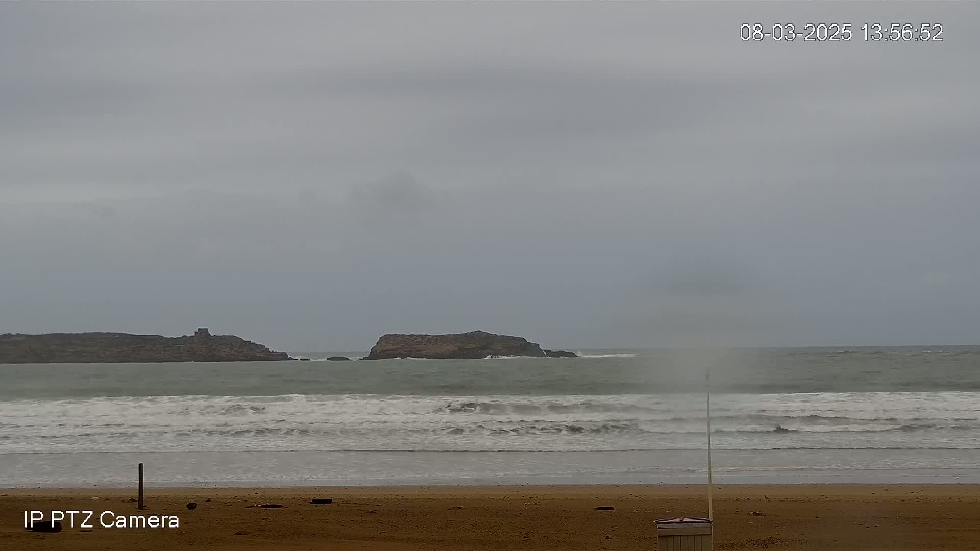 A sandy beach with moderate waves under an overcast sky shows a distant rocky island.