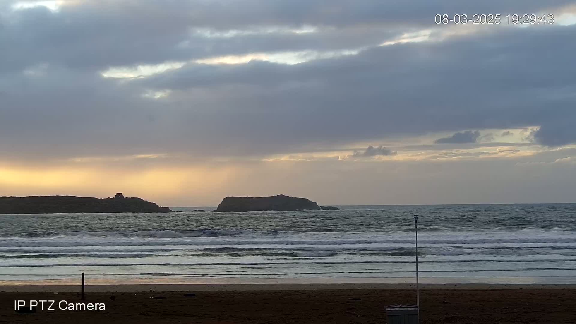 A sandy beach with moderate waves under a partly cloudy sky at sunset, with two small islands visible in the distance.