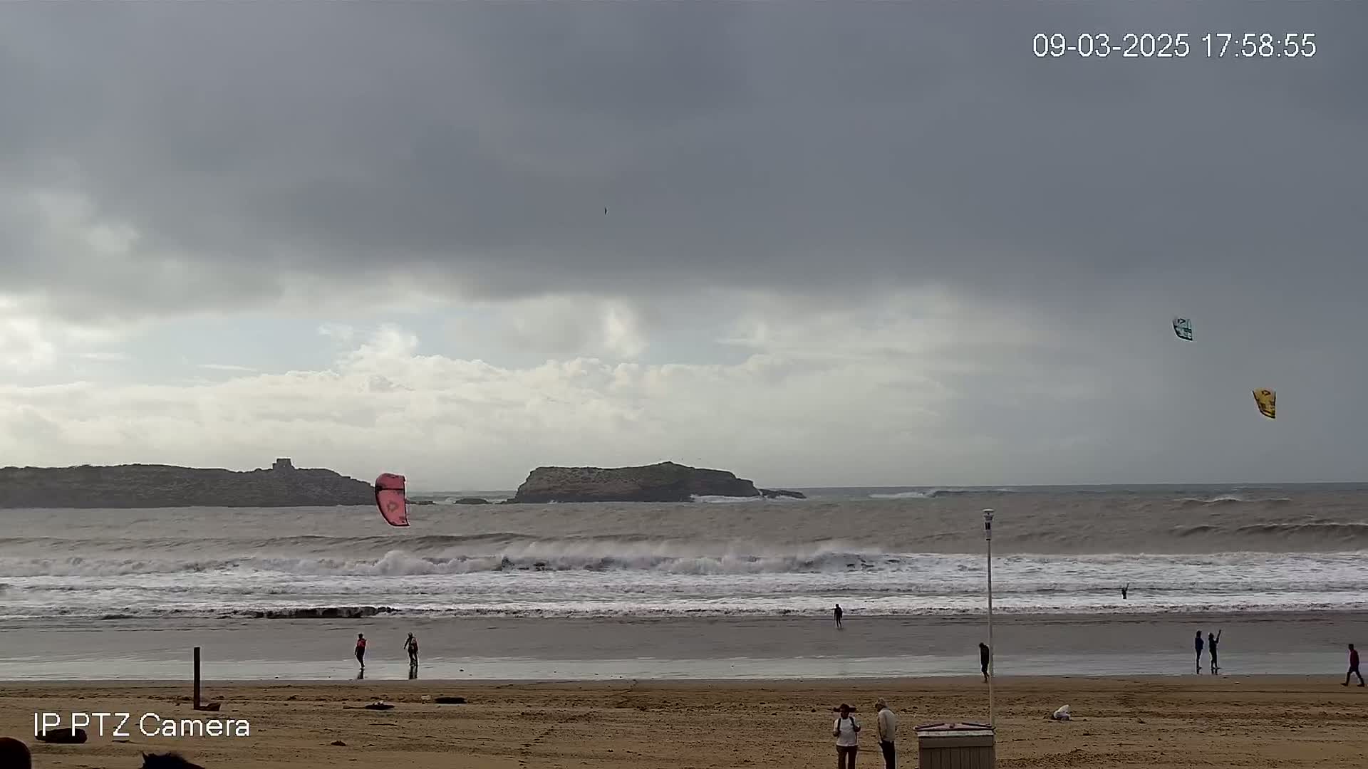 A cloudy, windy day at a beach with choppy waves, several people on the shore, and kitesurfers in the ocean.