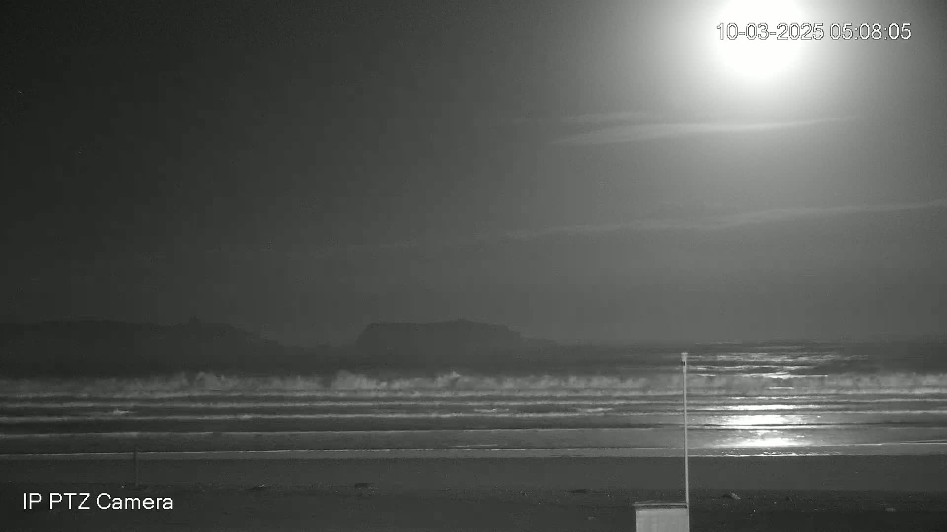 Under a bright moonlit night sky, waves crash gently onto a dark sandy beach, with a distant landmass visible in the background.