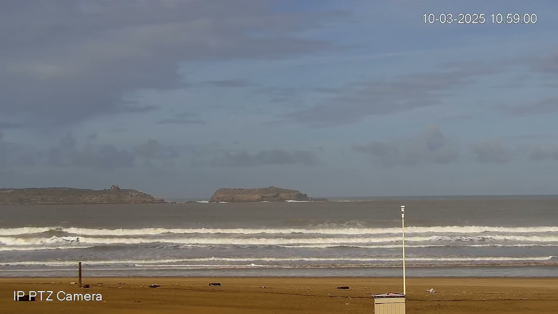 A sandy beach with small waves rolling onto the shore under a cloudy sky, with two small islands visible in the distance.