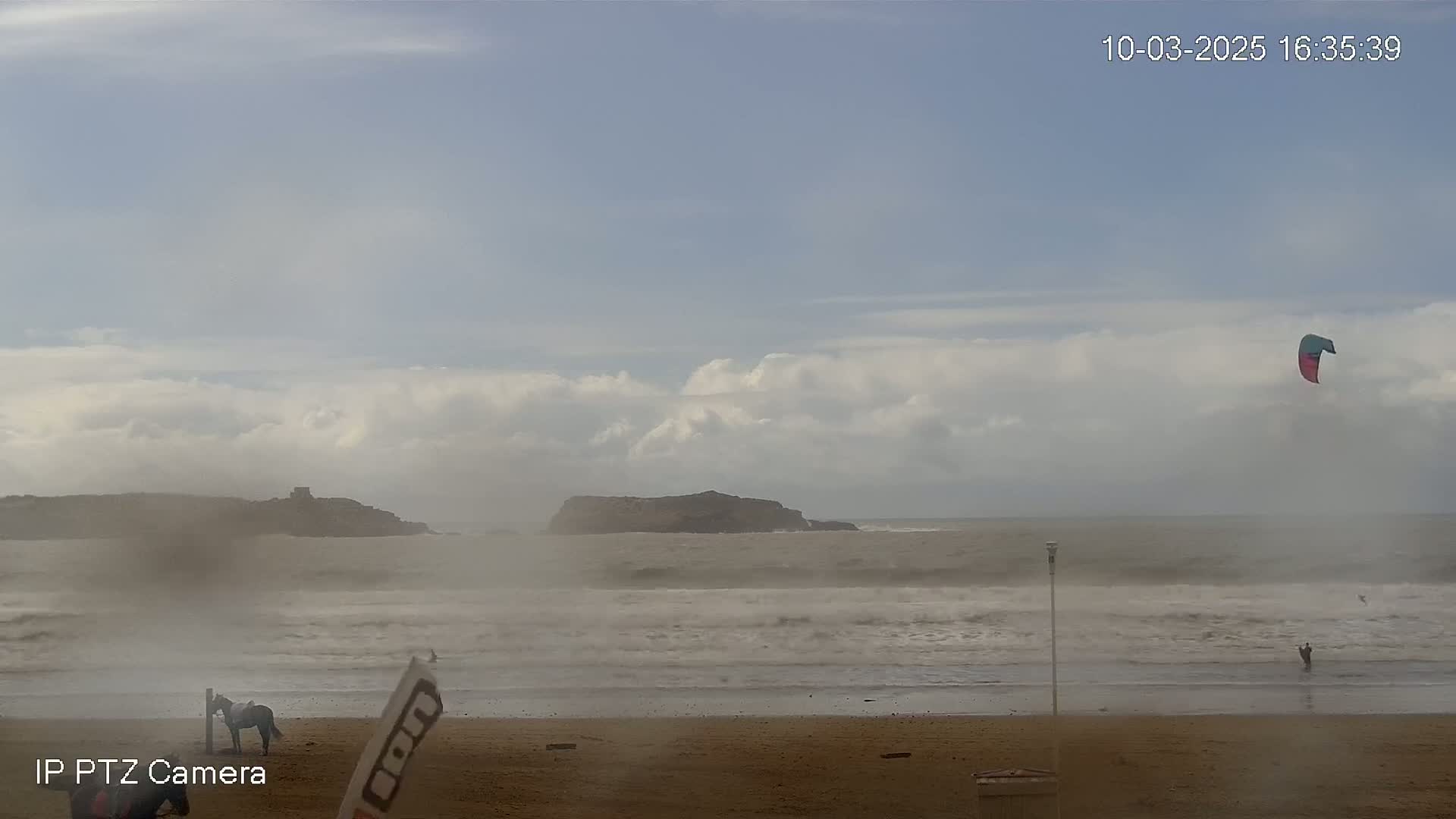 A sandy beach with a horse and rider, a kitesurfer in the water, and rocky outcrops in the distance under a mostly cloudy sky.