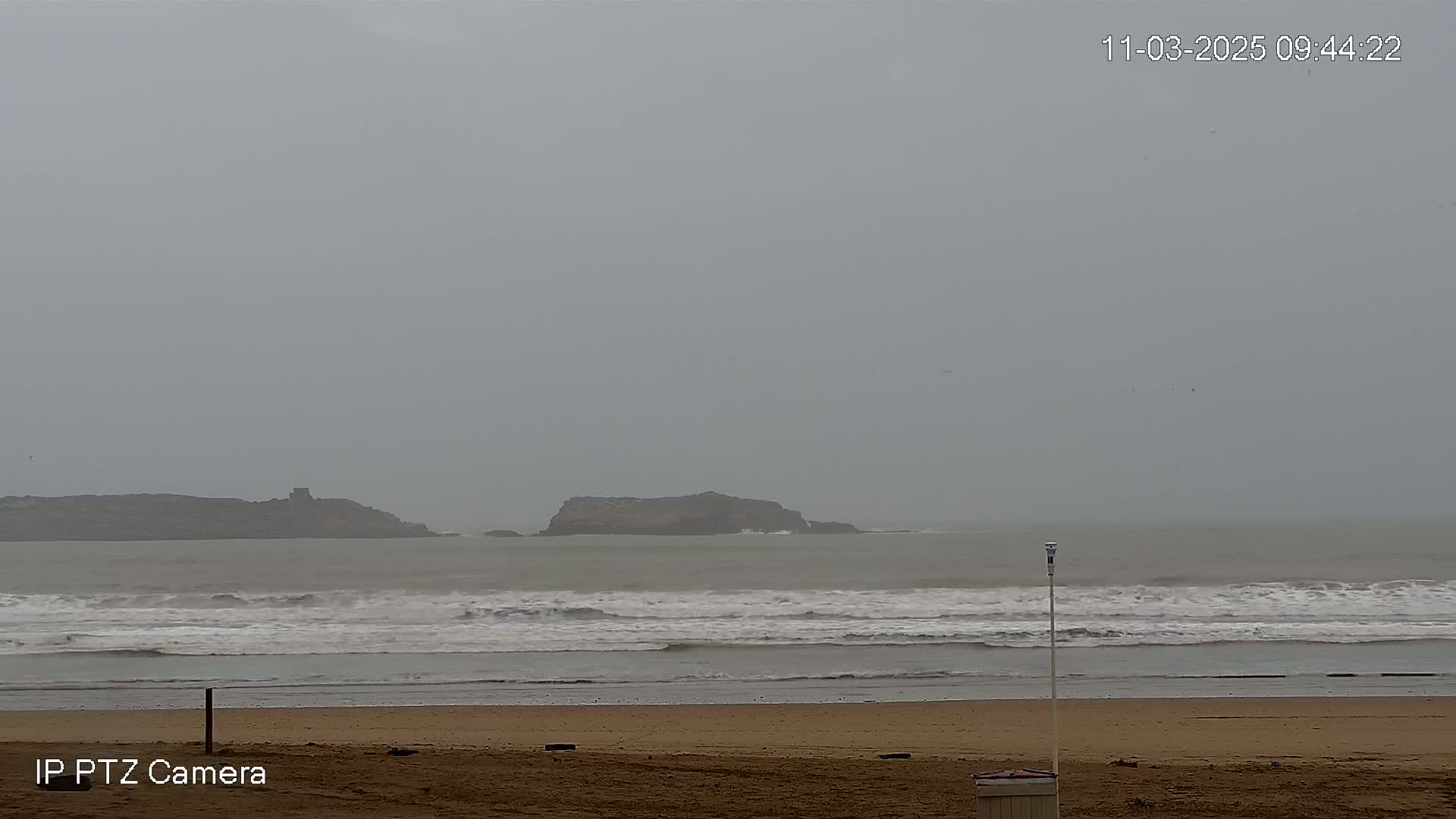 A sandy beach with moderate waves stretches to a distant rocky island under an overcast, gray sky.