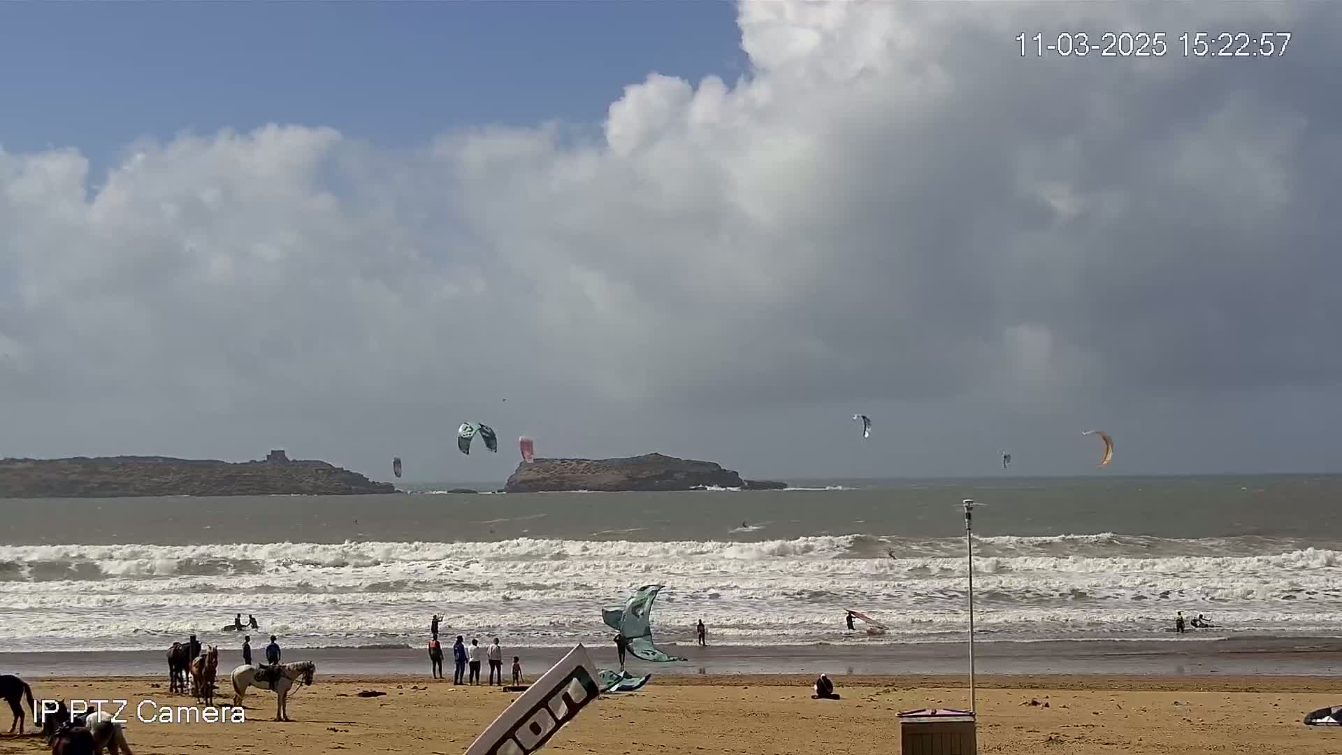 A sandy beach with several people, some riding horses, and numerous kitesurfers in the ocean under a partly cloudy sky.