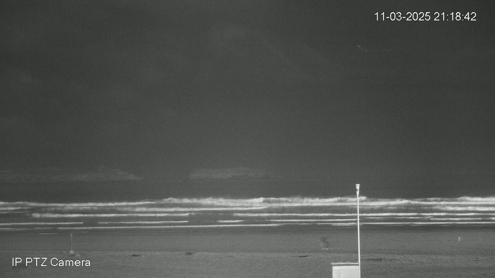 A nighttime, grayscale image shows a dark beach with gentle waves rolling in under a mostly clear sky.