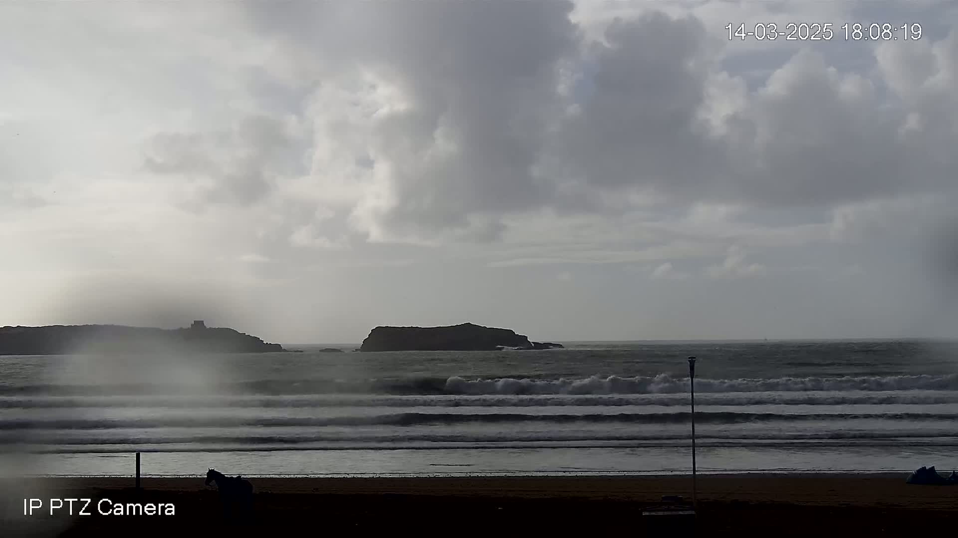 A mostly cloudy, overcast day shows a sandy beach with waves crashing on the shore, and two small islands visible in the distance.