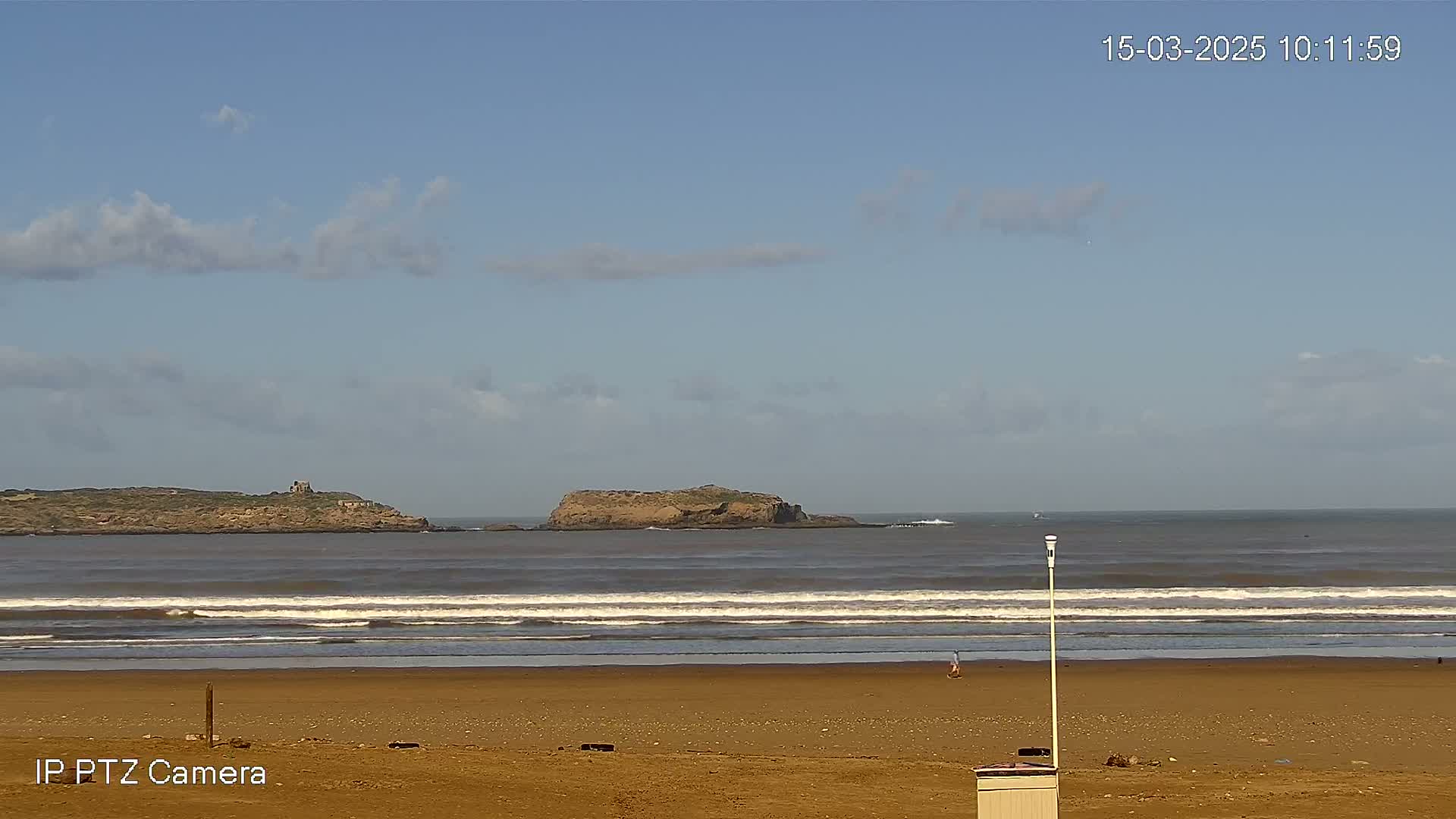 A sandy beach with small waves rolls onto the shore under a partly cloudy sky, with two islands visible in the distance.
