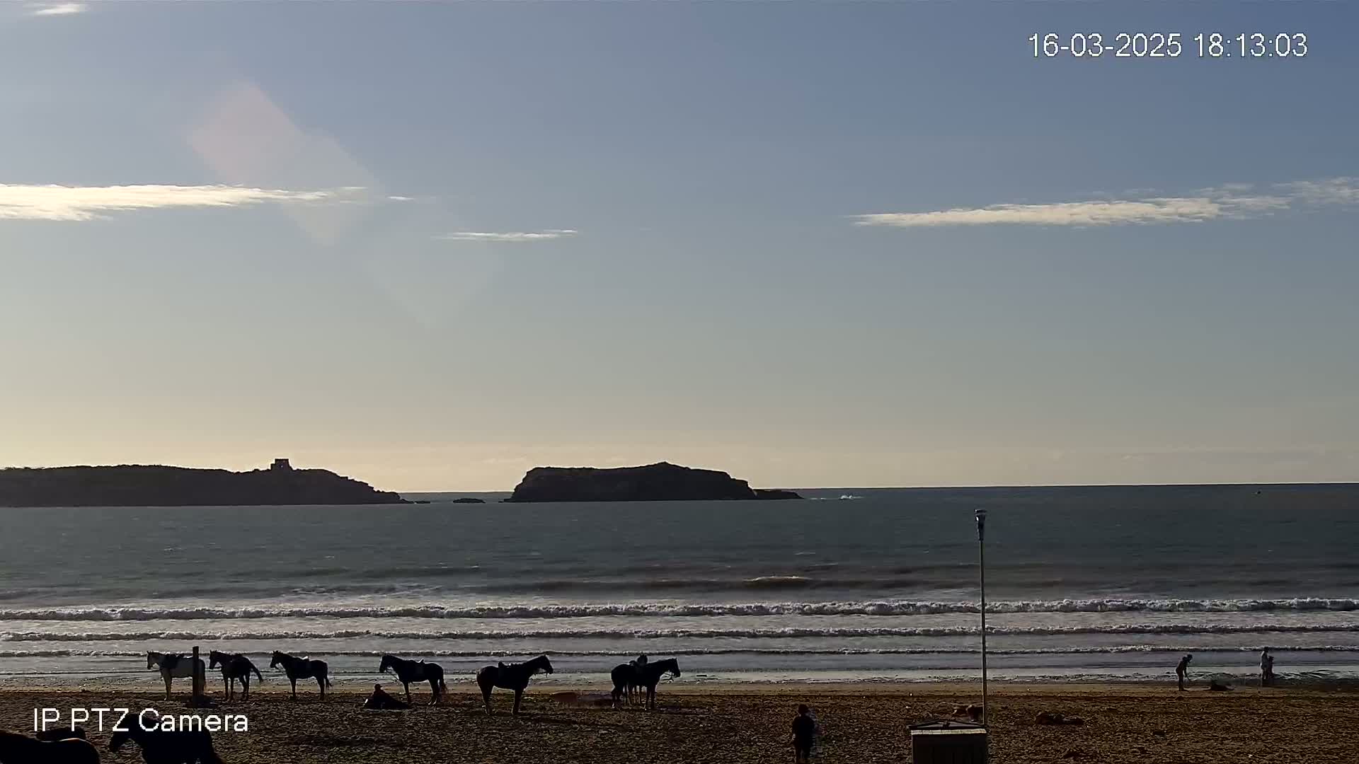 Several people are riding horses on a beach under a mostly clear sky, with a small island visible in the distance.
