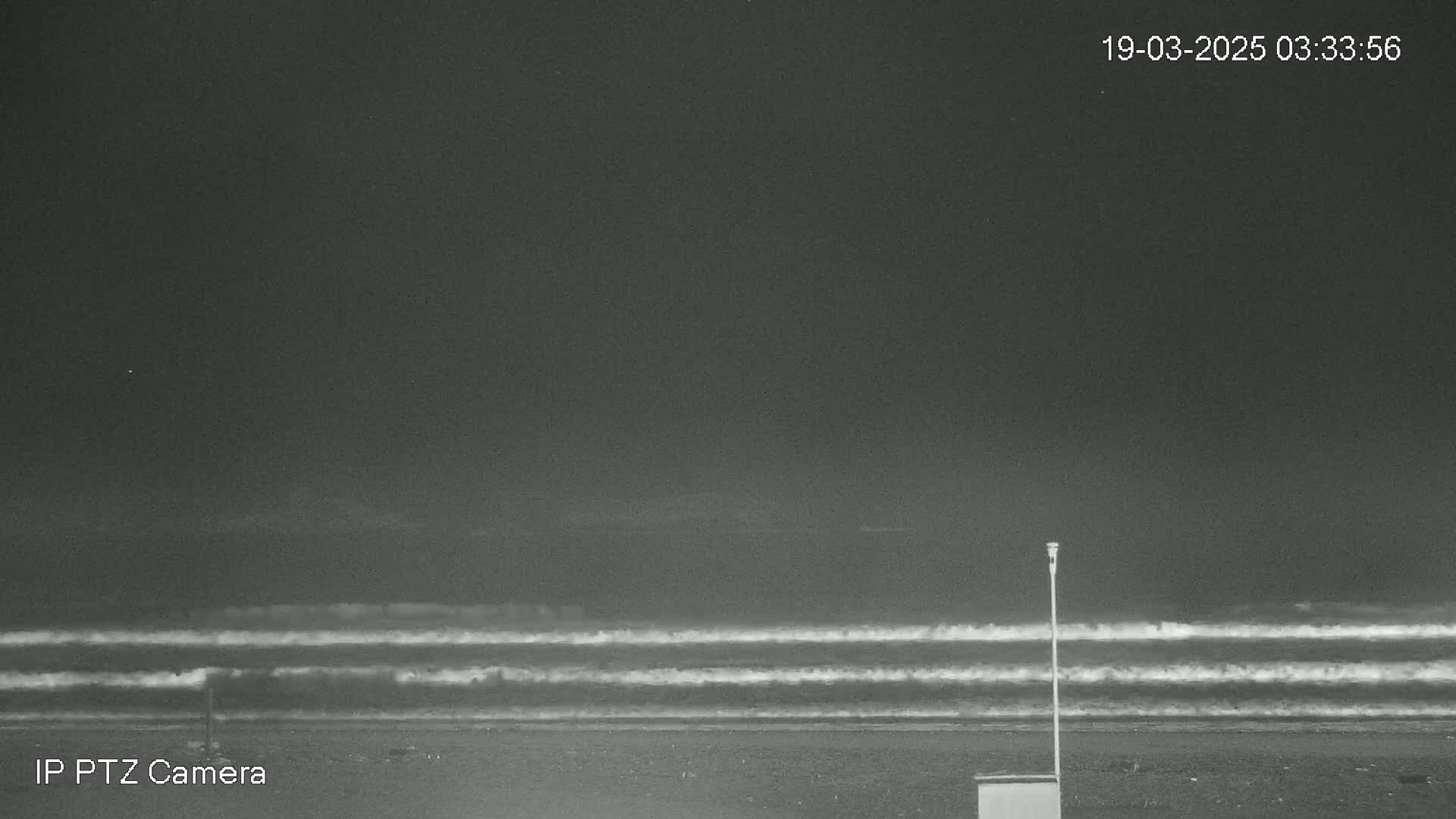 A dark, nighttime beach scene shows gentle waves rolling onto the shore under a cloudy sky.