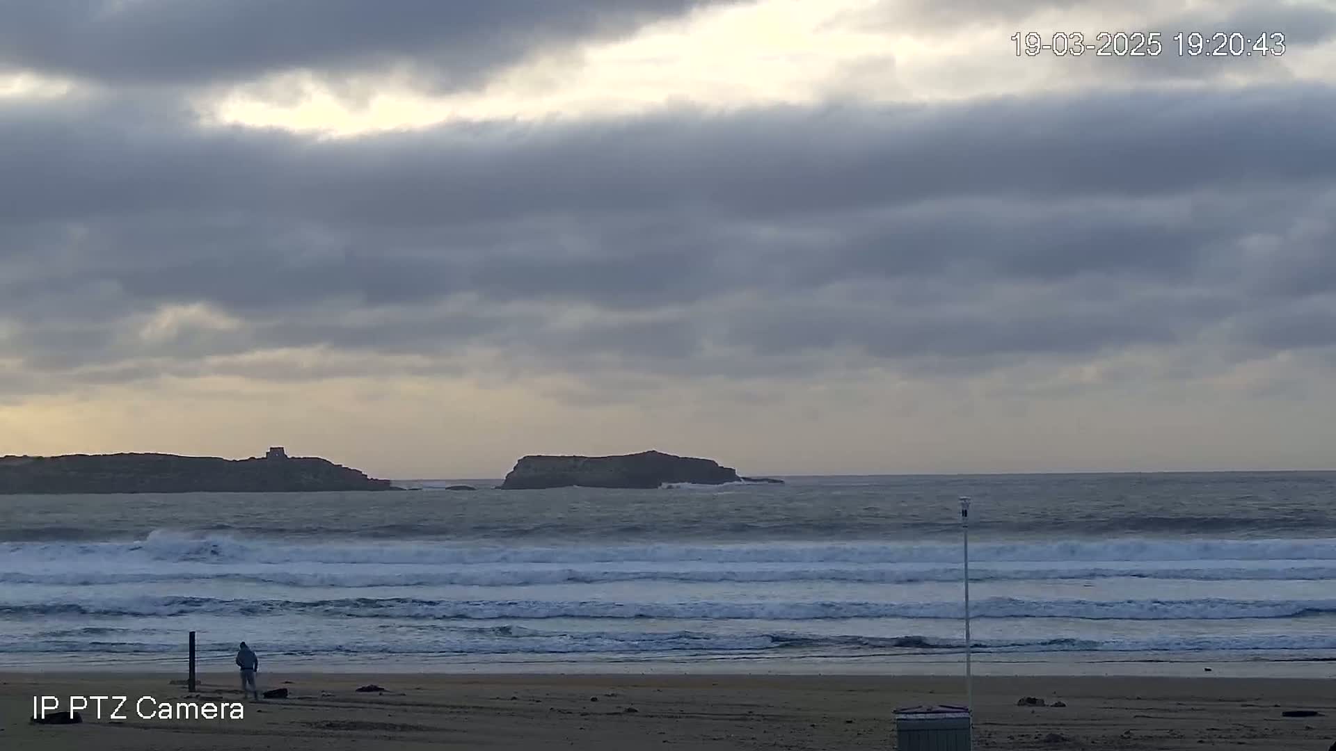A person stands on a sandy beach, watching waves roll in towards the shore under an overcast sky, with two small islands visible in the distance.