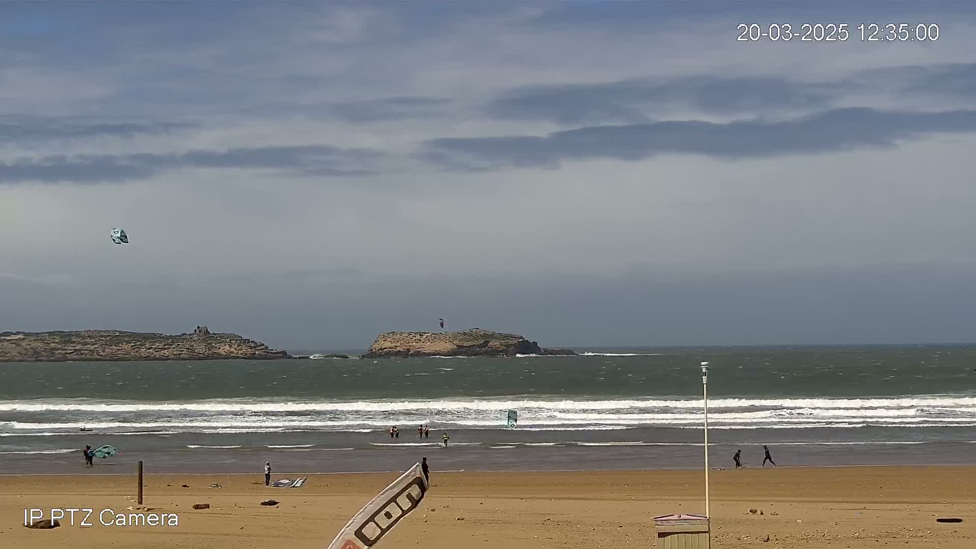 A sandy beach with several people and kitesurfing equipment, under a partly cloudy sky, overlooks a calm ocean with a small island in the distance.