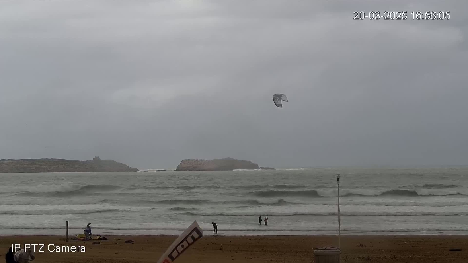 A kite surfer's kite floats in the overcast sky above a beach with several people on the shore and in the water, under a gray sky.