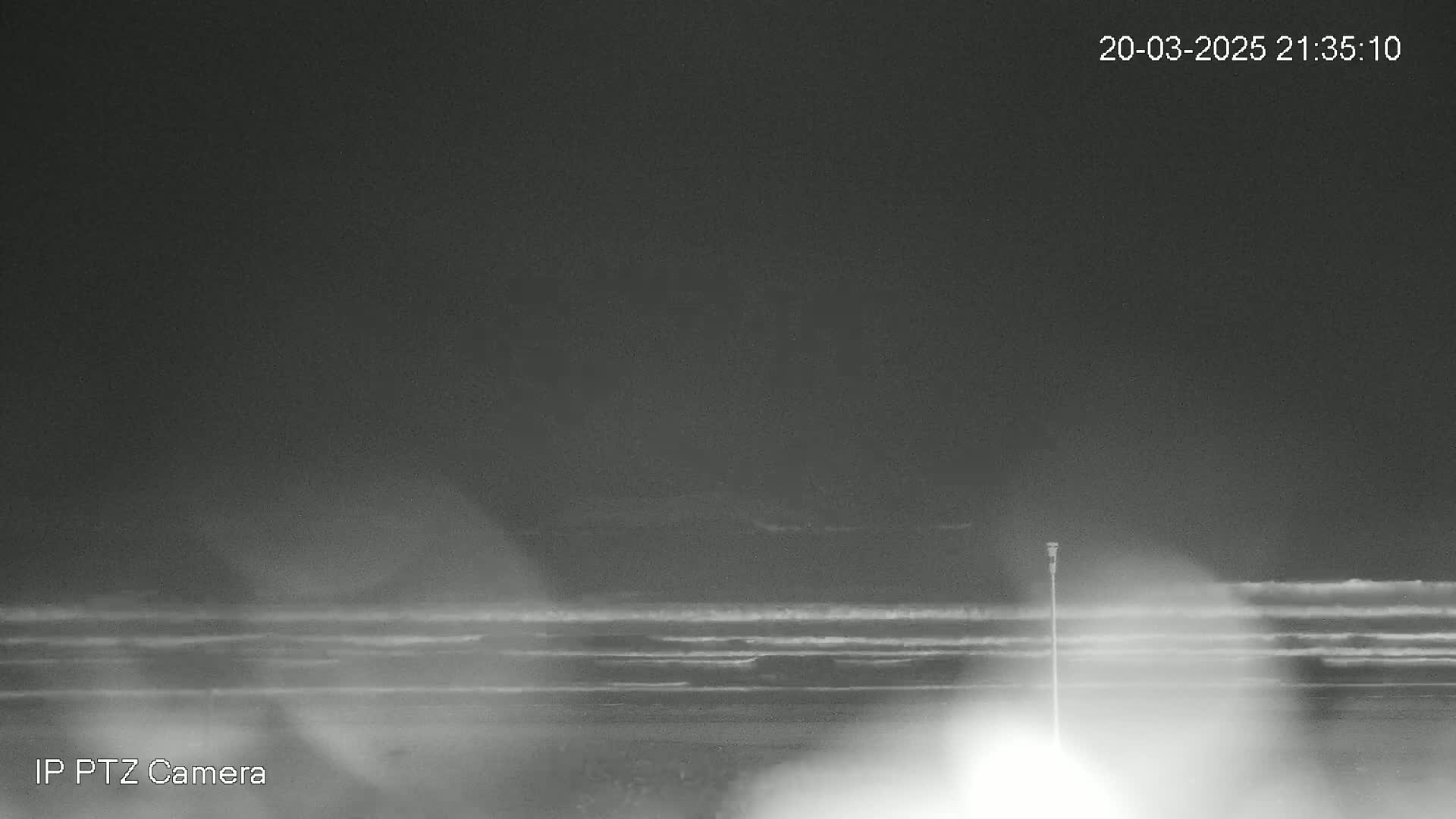 A nighttime, low-light image shows a dark beach with gently rolling waves under a cloudy sky.