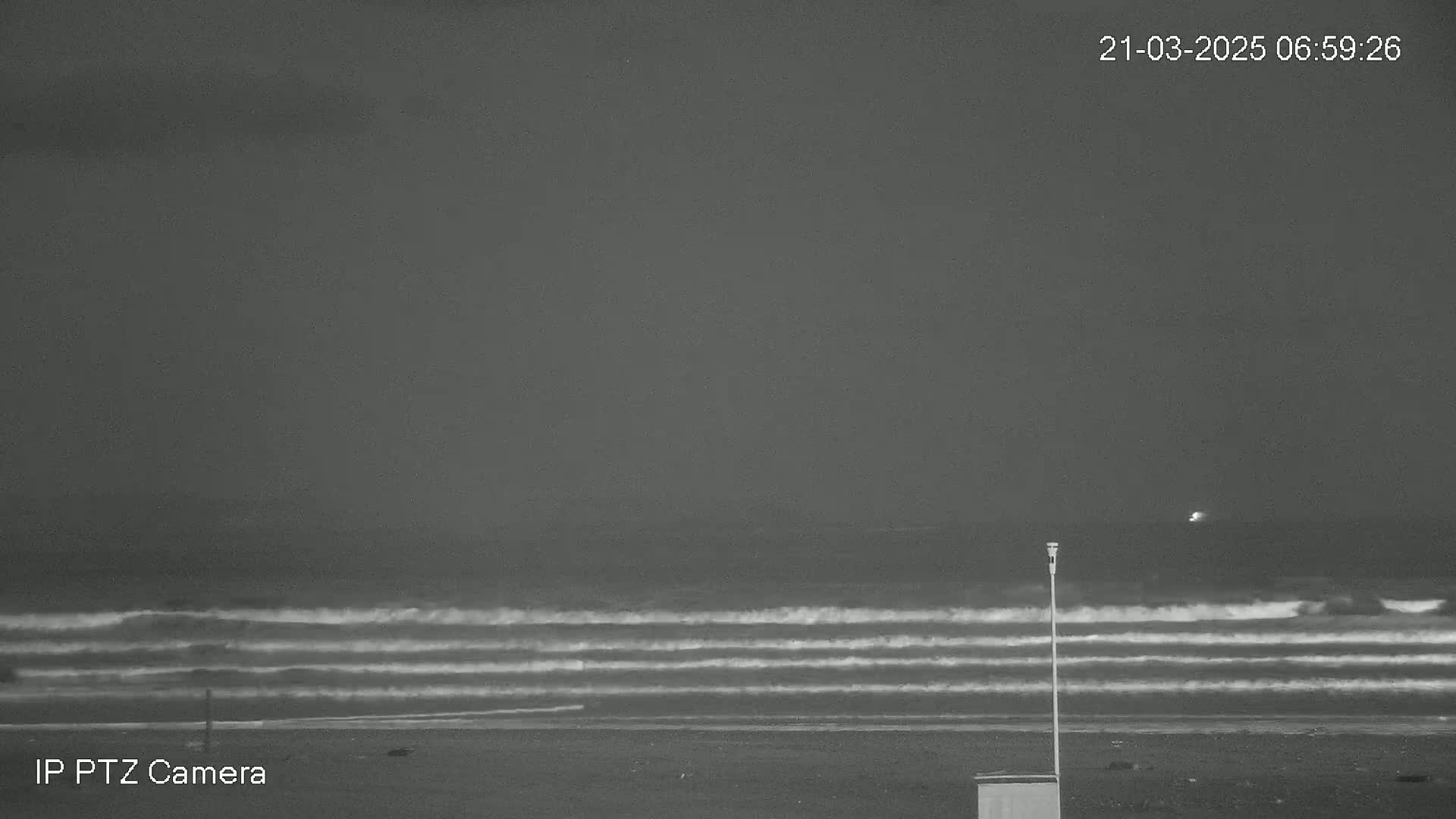 A dark, nighttime beach scene shows a relatively calm sea with small waves rolling onto the shore under a gray sky.