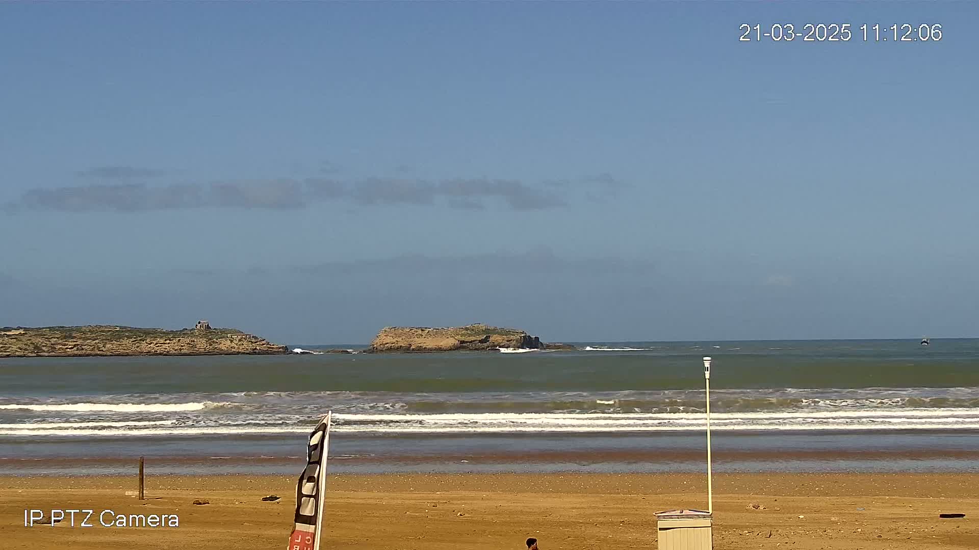 A sandy beach with gentle waves laps the shore under a mostly clear blue sky, with two small islands visible in the distance.