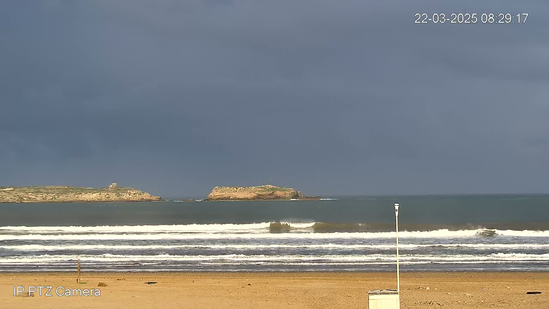 A sandy beach with gentle waves rolls onto the shore under a dark, overcast sky, with a small island visible in the distance.